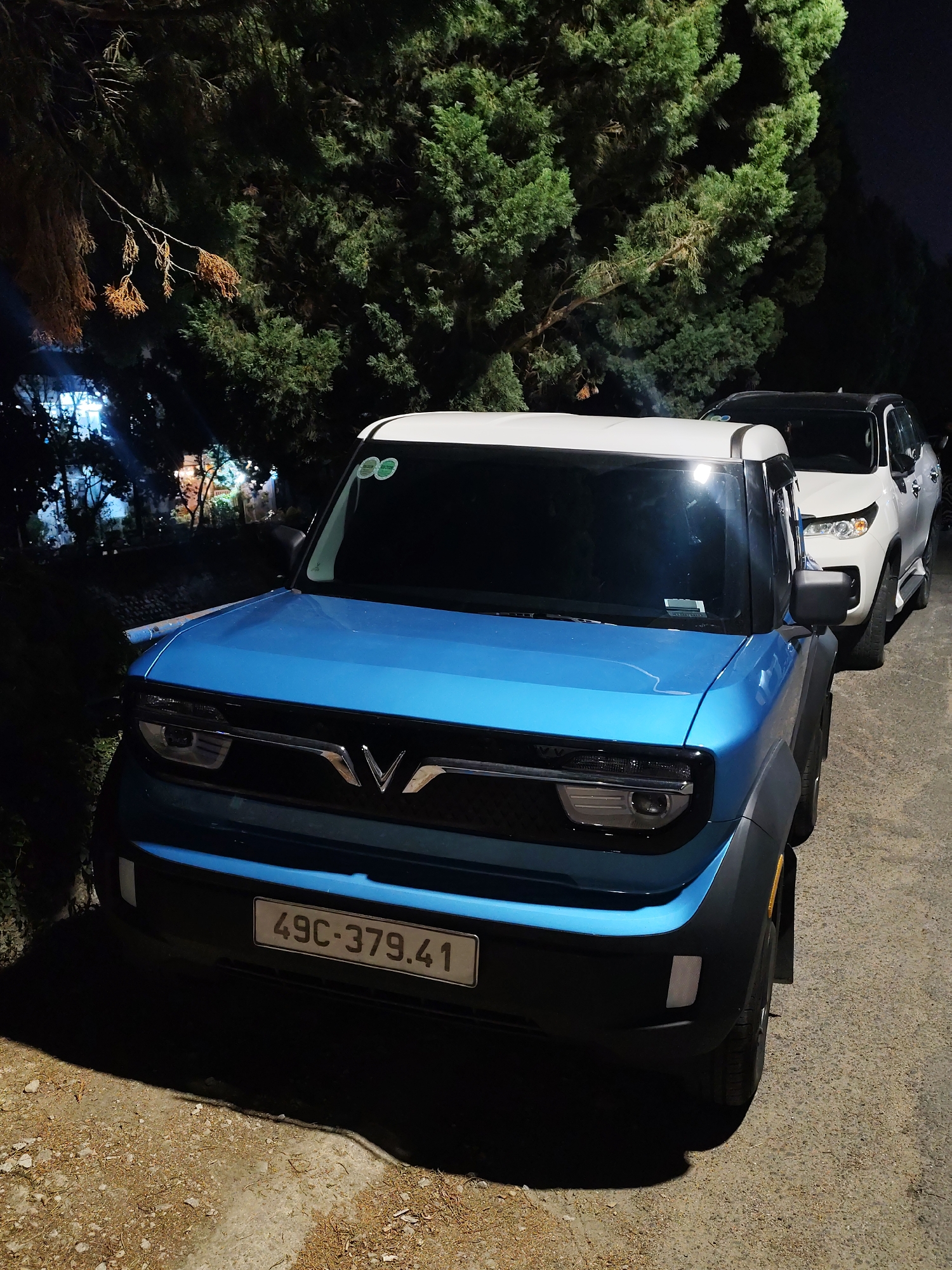 The image presents a nighttime scene in Da Lat, Vietnam, featuring two parked vehicles against a backdrop of dense green trees. In the foreground, a blue and white VinFast VF 3 electric car is prominently positioned, angled slightly towards the viewer. The car's body is a vibrant blue, complemented by a white roof. The distinctive "V" logo of VinFast is visible on its front grille. A Vietnamese license plate, "49C-379.41," is clearly displayed on the front bumper. Small, circular green stickers are also visible on the top right portion of the windshield.
Behind the VinFast VF 3, a white SUV, appearing to be a Toyota Fortuner, is parked, with its front section partially visible. Both vehicles are situated on a rough, possibly gravel or poorly paved, ground surface, which is unevenly illuminated by artificial light sources.
The setting is dominated by a thick cluster of dark evergreen trees, likely conifers, forming a dense canopy that fills the background and frames the cars. Some dry, brown branches hang from the trees on the left side, adding textural contrast. Through the gaps in the foliage on the far left, distant artificial lights from buildings or structures are discernible, indicating a nearby developed area. The overall ambiance is one of quietude in a natural or semi-natural environment after dark. No people are visible, and the cars appear to be stationary. The dim, artificial lighting casts long shadows and highlights specific features of the cars and ground, leaving much of the surrounding natural elements in deep shadow.