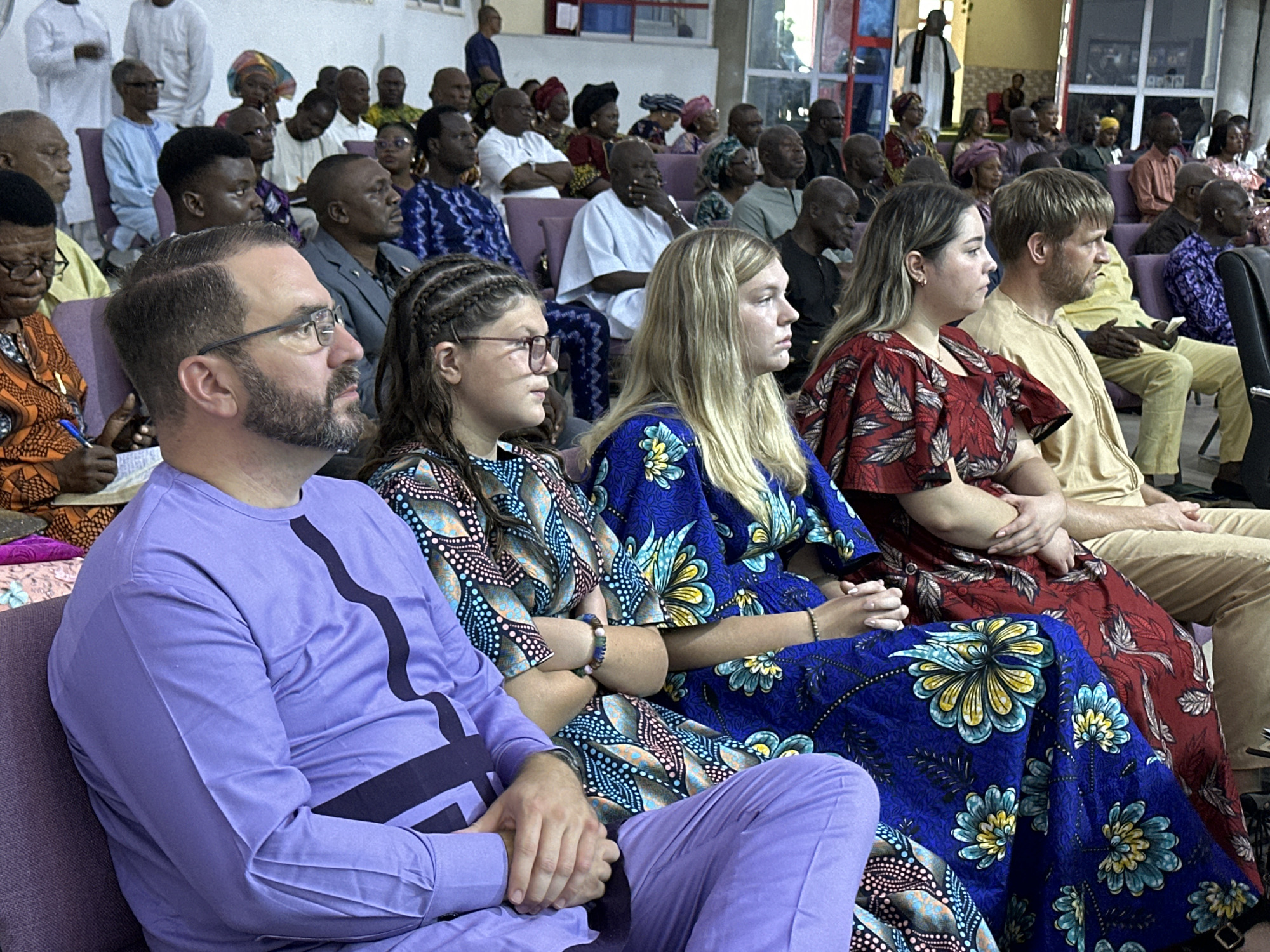 In Television, Nigeria, a group of people are seated in a room, possibly a church or community hall, looking towards the front. The focus is on a seated group in the foreground, with a man in a lavender outfit on the left, and two women in patterned dresses to his right. The man has a beard and glasses, and his hands are clasped in his lap. The woman closest to him has braided hair and is looking forward. The woman next to her has blonde hair and is also looking forward, with her arms crossed. Behind them, other individuals are visible, many in colorful African attire. The room appears to be a place of worship or a gathering space, with rows of seating and some architectural elements visible in the background. The lighting suggests an indoor setting.