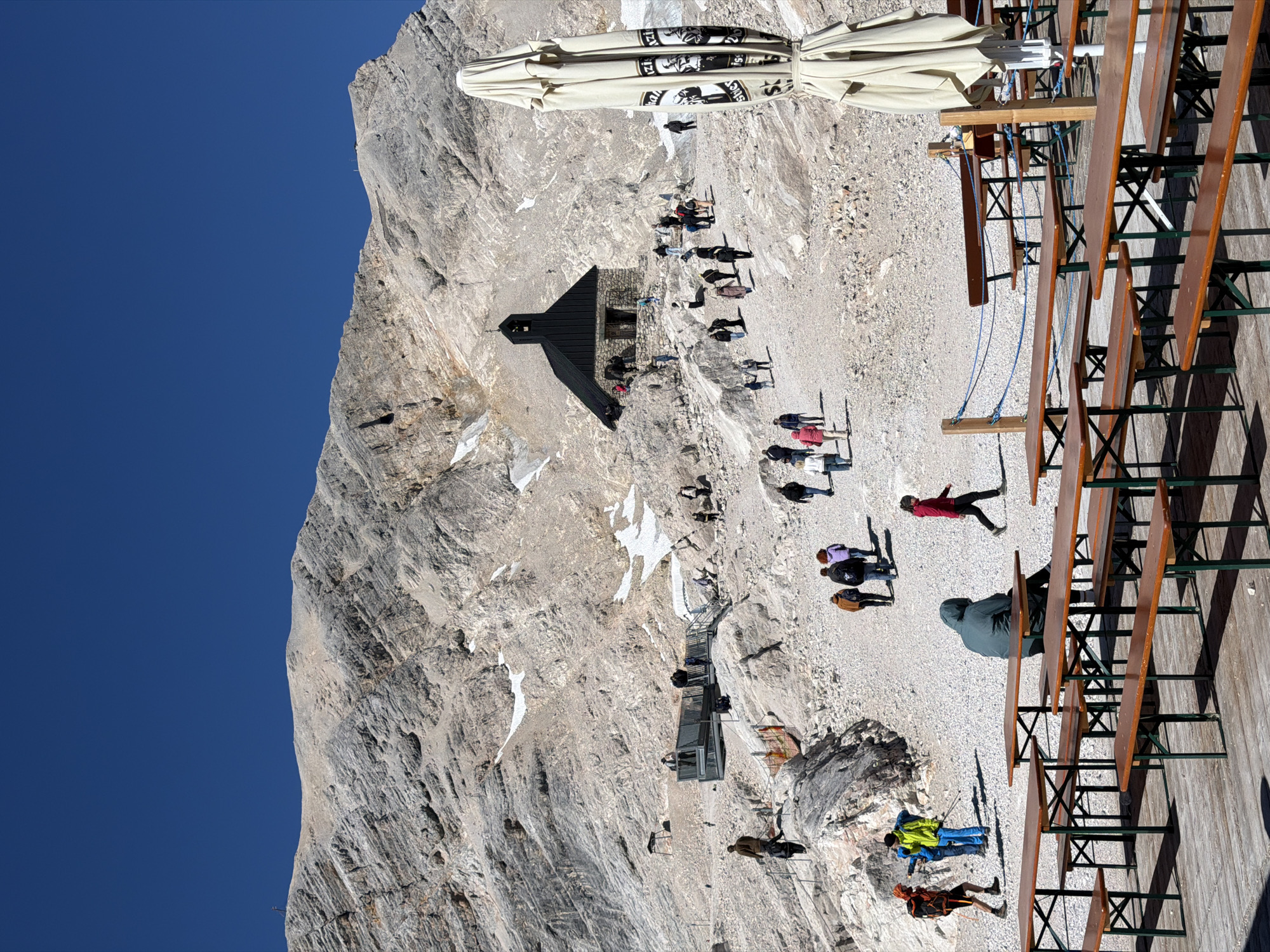 This image depicts the Zugspitze, Germany's highest peak, located near Garmisch-Partenkirchen. The scene is characterized by a steep, rocky mountainside under a clear blue sky. In the mid-ground, a small stone chapel with a dark roof and a bell tower stands on a gravelly slope. A group of tourists are walking up a path towards the chapel.
In the foreground, numerous wooden picnic tables and benches are set up on a wooden deck, suggesting an outdoor seating area for a restaurant or cafe. One person is seated at a table, wearing a hooded green jacket. To the right, a large, folded beige umbrella with black lettering stands near the tables.
Further back and to the left, a modern elevated walkway or viewing platform with railings is visible, with people standing on it. The surrounding terrain is rugged and rocky, with patches of snow clinging to shaded areas of the mountainside. Several individuals are scattered across the landscape, dressed in outdoor clothing suitable for a mountainous environment. The overall impression is a popular tourist destination in a high alpine setting.
