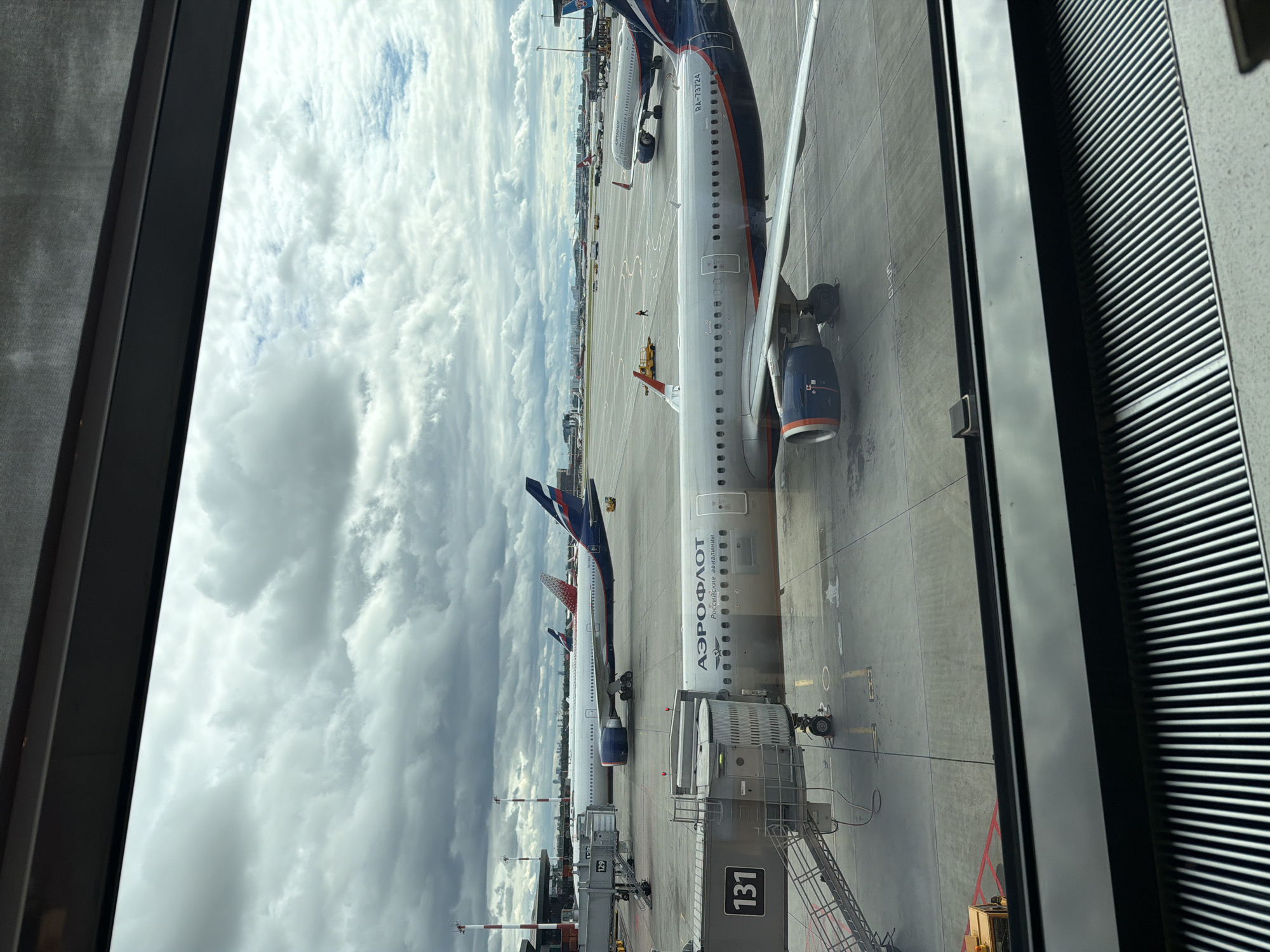 The image depicts an airport tarmac with multiple commercial airplanes. In the foreground, an Aeroflot airplane is parked at gate 131, connected to a jet bridge. Another Aeroflot aircraft is visible behind it, with its tail fin featuring the Aeroflot logo. Further in the background, other airplanes are seen on the tarmac, with buildings and infrastructure typical of an airport. The sky is overcast with clouds. The location is Sheremetyevo International Airport (SVO) in Khimki, Russia, as evidenced by the Aeroflot branding and the airport's layout. No people are clearly identifiable in the image, and the scene appears to be in a state of operational normalcy for an airport.