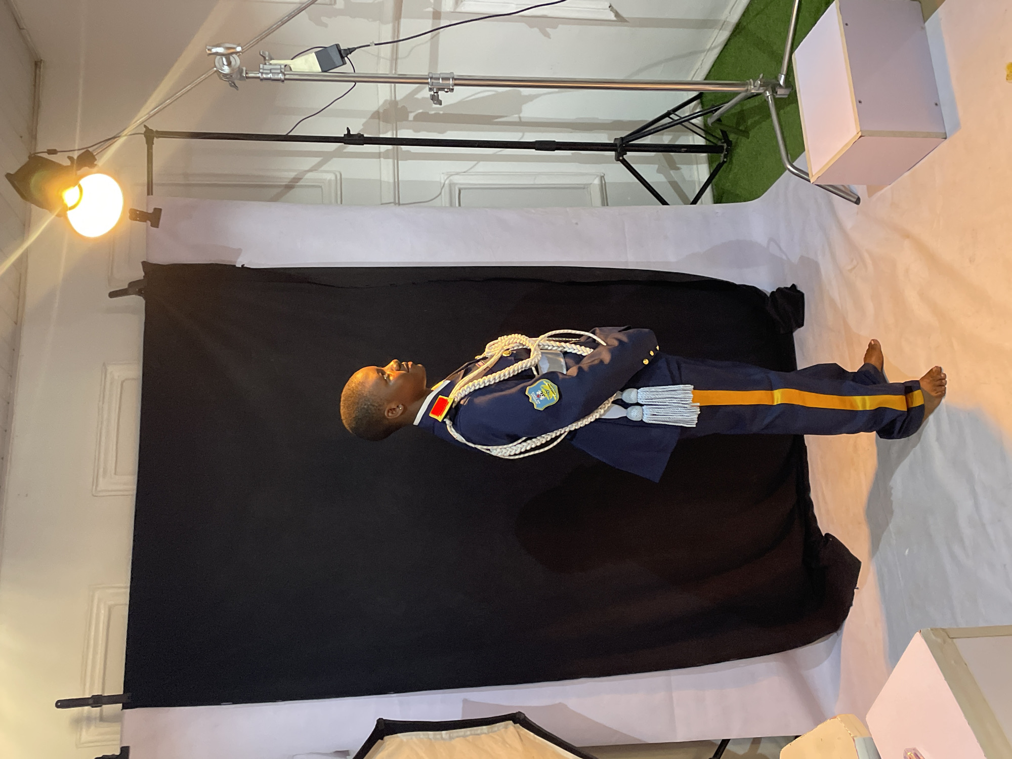 A studio portrait of a young man in a military-style uniform with blue and gold trim. The uniform has a white rope detail and a badge. He is lying on his side against a black backdrop, which is part of a studio setup with lighting equipment visible. The floor is covered with white fabric or paper, and a white box-like structure is on the right side of the frame. The location is a photography studio, presumed to be in Barakin Akawo, Nigeria. The setting is indoors with artificial lighting. The man is barefoot.