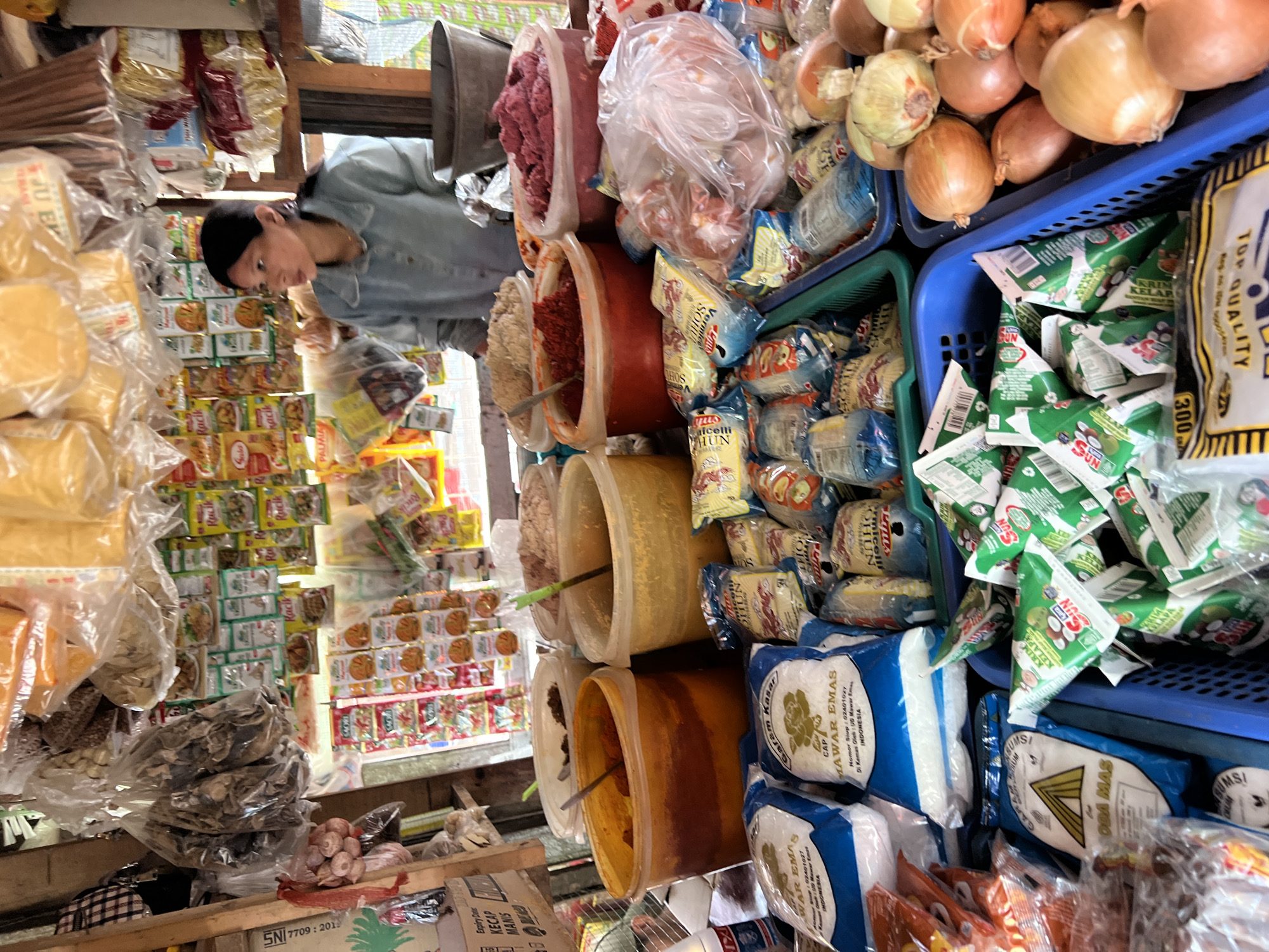 The image shows a street food stall in Citeureup, Indonesia, filled with various food items. A young woman is standing behind the counter, wearing a light blue shirt. She appears to be a vendor. The stall is packed with sacks of rice, bags of spices, and containers of condiments. In the foreground, there are blue and white sacks of what appears to be rice, along with other packaged goods. Behind the woman, shelves are stocked with various food products, including small packets of spices and larger bags of yellow and orange powders. There are also large plastic buckets filled with different food ingredients. The overall impression is of a bustling local market stall, offering a wide variety of food staples and spices to customers.