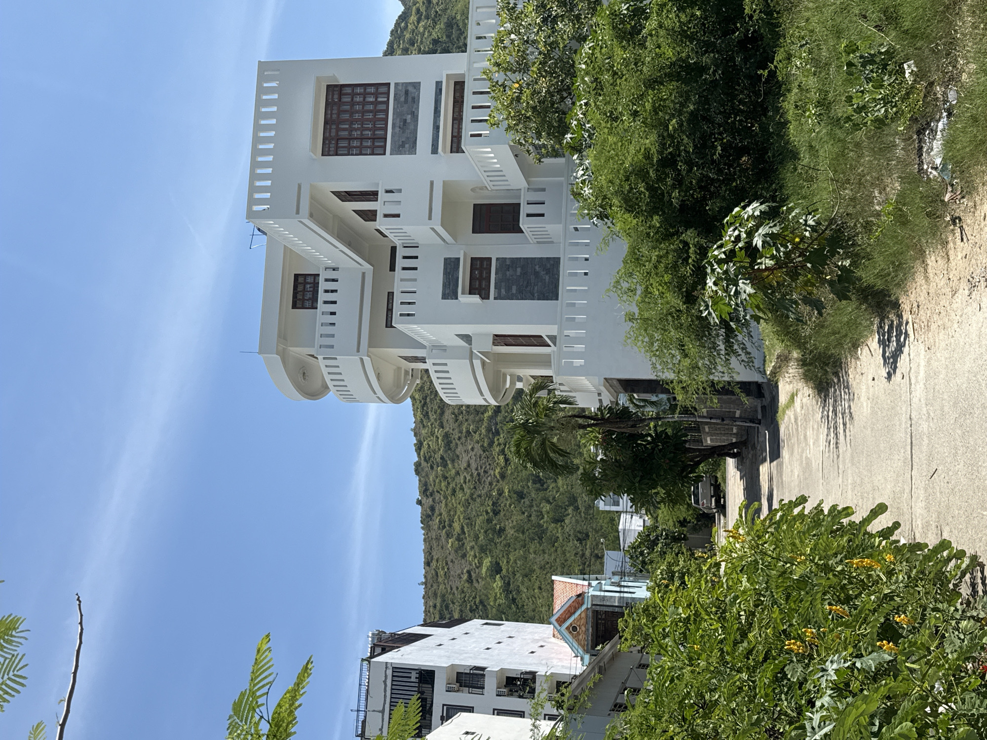 A residential street in Bac Nha Trang Ward, Vietnam, is shown on a sunny day under a bright blue sky with wispy clouds. In the foreground on the left, lush green bushes with yellow flowers line a concrete path that recedes into the distance. The path is bordered by a grassy verge on the right.
Dominating the right side of the image is a multi-story white building with a modern, angular design. It features numerous balconies with white railings, dark-framed windows, and decorative grey stone accents. To the left of this building, other residential structures are visible, including a taller, white building with balconies and several smaller houses with pitched roofs.
Further down the street, a silver SUV is parked. The background consists of a densely vegetated hillside that rises steeply behind the houses. The overall scene depicts a quiet, sun-drenched neighborhood with a mix of modern and traditional architecture, surrounded by abundant greenery.