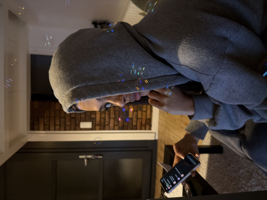 A person in a gray hoodie looks down at a smartphone held in their right hand. The background shows a modern interior with brick accents, dark cabinetry, and white walls. Bubbles float in the air, some reflecting colorful light. The image is rotated clockwise by 90 degrees.