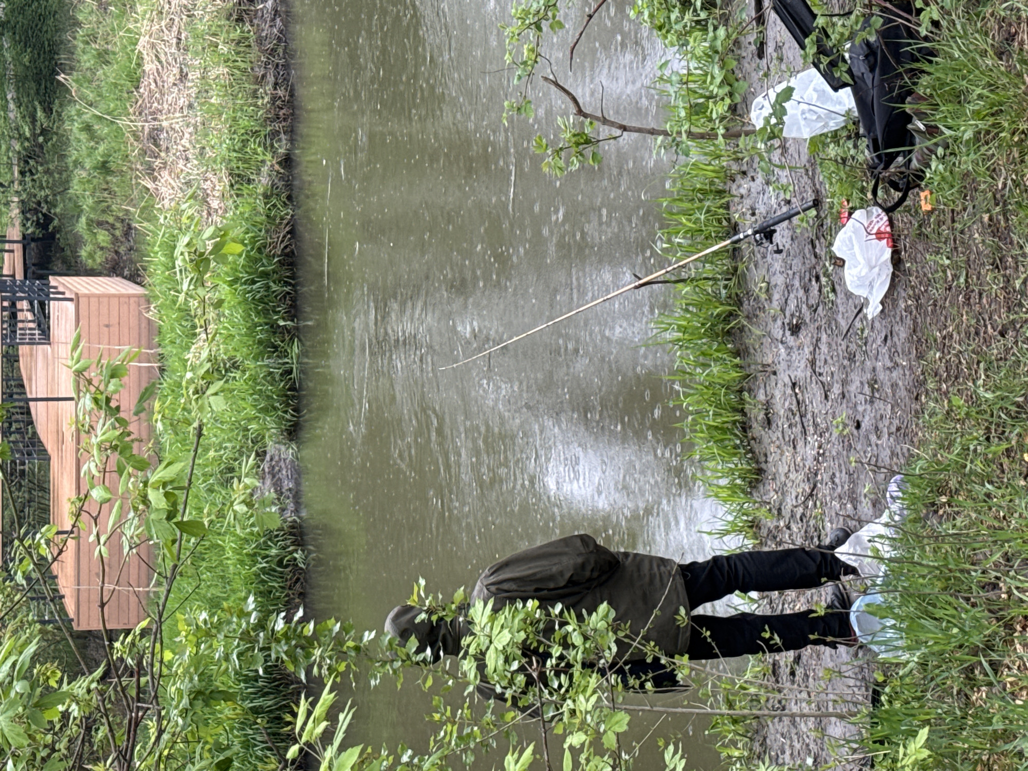 A man wearing a dark green jacket and black pants stands on the muddy bank of a body of water, possibly a pond or small river, near what appears to be a wooden structure or platform. He faces the water. A fishing rod is positioned nearby, extending out over the water. White plastic bags and a dark backpack are also on the bank, near the rod. The surrounding environment is lush with green vegetation, including grass, bushes, and trees. The water is murky, reflecting the sky and surrounding foliage. The location is Moscow, Russia.
