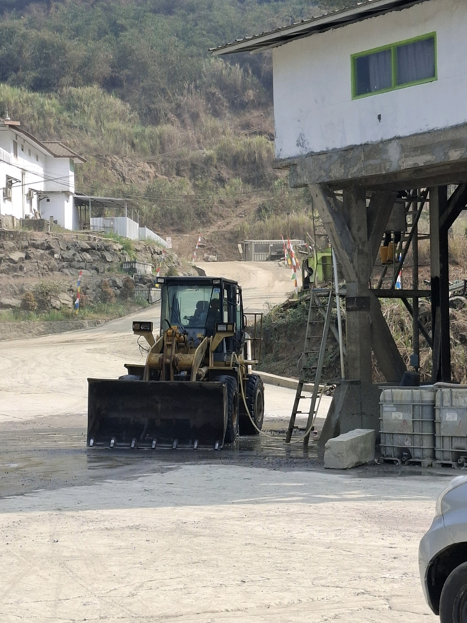 The image depicts a construction site in Cijangkar, Indonesia, featuring a yellow front-end loader parked on a dusty, wet surface. The loader has its bucket lowered to the ground. In the background, a white building with a green-framed window is visible, elevated on concrete stilts. A steep, vegetated hillside rises behind the structures. A metal ladder leans against the stilts of the building, and several large industrial tanks are situated nearby. The overall environment suggests an active work area with ongoing construction or material processing.