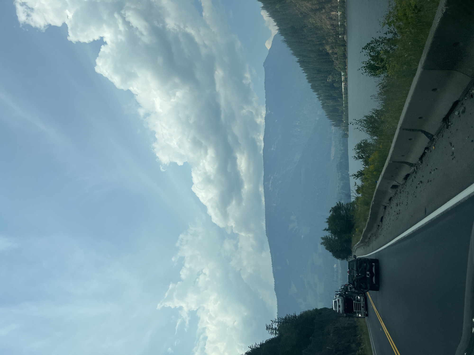 The image depicts a scenic view of a highway alongside a body of water and mountainous terrain. On the road, a large truck carrying multiple vehicles is traveling in the left lane, followed by a dark pickup truck in the right lane. The road is paved and bordered by a concrete barrier on the right. The landscape features a dense forest covering the slopes of the mountains, which rise in the background. A large, calm body of water is visible to the right of the road, reflecting the sky. The sky is partly cloudy, with patches of blue sky interspersed with large, white cumulus clouds. The overall impression is that of a natural, open-air environment, likely a highway in a mountainous region. The location is specified as Area E (Eagle River Valley), Canada.