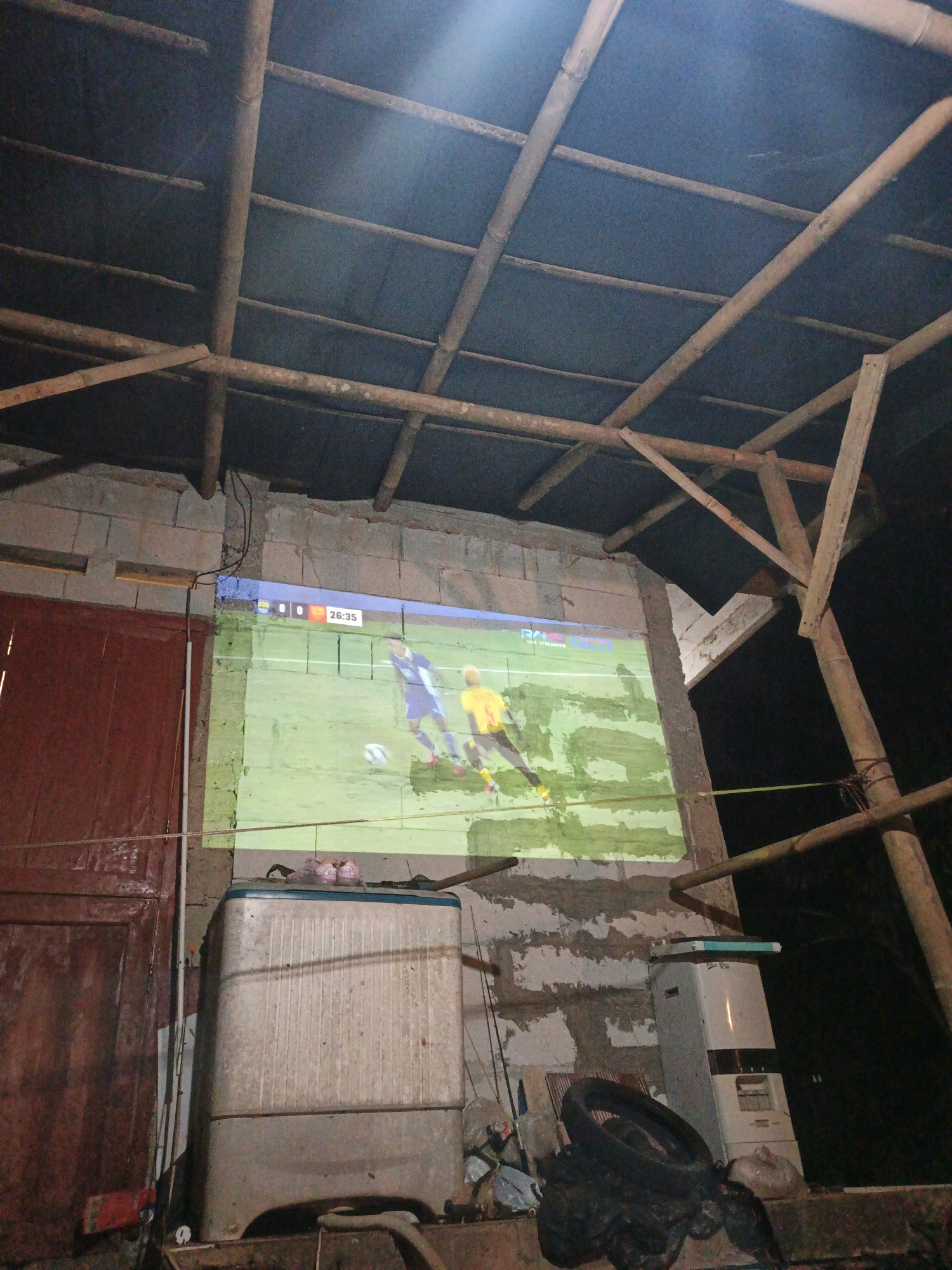 This is a medium-shot, eye-level, natural light photograph taken at night in an outdoor setting that appears to be a residential or makeshift outdoor viewing area. The scene depicts a projector screen set up against a rough, unpainted concrete block wall. The screen displays a live broadcast of a soccer match, with two players in blue and yellow jerseys visible on a green field. The score "0 0" and the time "26:35" are displayed in the upper corner of the screen, indicating the game is in progress.
The immediate foreground shows a dirty, old-fashioned washing machine with a blue trim, positioned to the left of the projector screen. Above the washing machine, on the wall, a pair of pink flip-flops are visible. To the left of the washing machine, there is a reddish-brown wooden door.
The background reveals a rustic roof structure made of bamboo poles and dark-colored sheeting. In the lower right portion of the image, a discarded tire is seen, along with various plastic bags and other debris, suggesting a less-than-pristine environment.
The overall impression is of an informal, possibly improvised setup for watching television or a sporting event outdoors. The environment suggests a developing or rural area, consistent with a location in Tasikmalaya, Indonesia, where such resourceful setups are common. The lighting is primarily from the projected image, with some ambient light contributing to the scene. The scene is static, with no active interactions observed, beyond the broadcast itself.