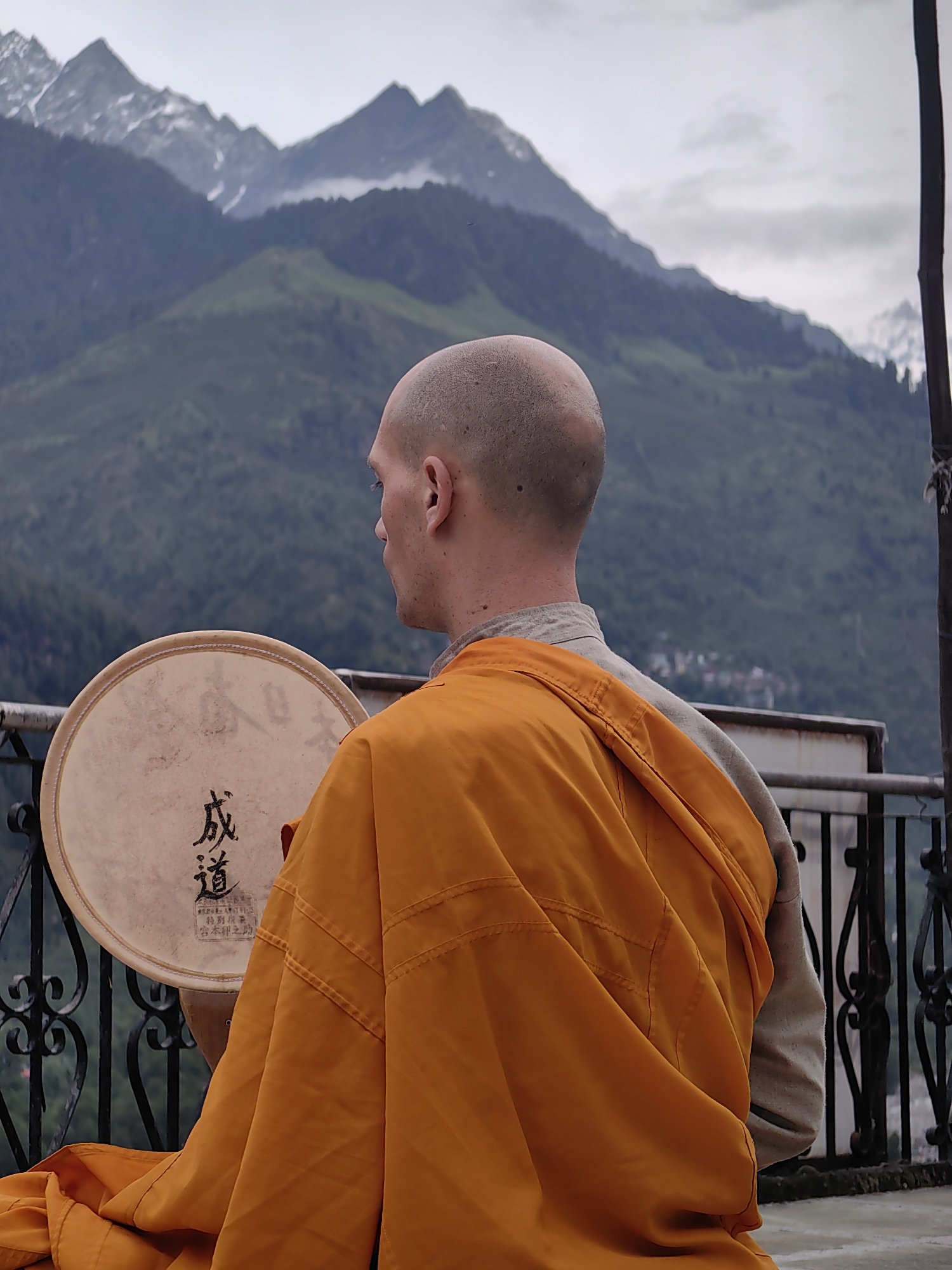A bald man, wearing an orange robe over a grey garment, sits outdoors with his back to the camera, holding a drum. The drum is light brown with black characters written on it. The man is positioned on a balcony with a black metal railing. In the background, a village is visible nestled on a green mountainside, with snow-capped mountains rising behind it under a cloudy sky. The location is identified as Vashisht, India.