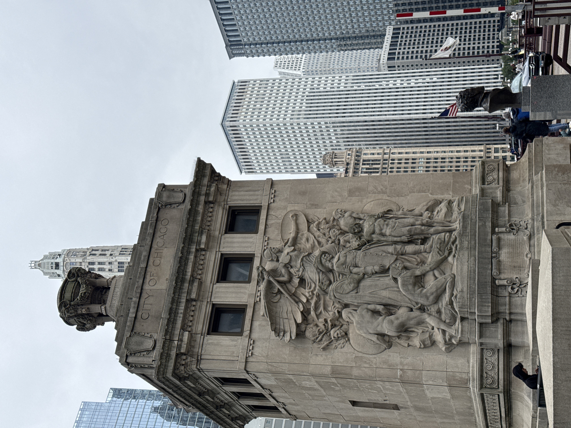 This is a vertical, eye-level shot of a stone sculpture adorning a building in Chicago. The sculpture depicts a mythological or allegorical scene with multiple figures, including what appears to be an angel with wings. The stone is weathered and detailed, with figures in dynamic poses. The building itself is constructed of stone and features a repeating pattern of windows. The words "CITY OF CHICAGO" are carved into the stone above the sculpture. In the background, several modern skyscrapers with glass facades rise into a cloudy sky. The lighting suggests it is daytime, but the overcast weather creates a muted, cool tone. A few people are visible in the distance, adding a sense of scale to the monumental architecture. One person is leaning out from a railing, looking at the sculpture. Another person in a dark hoodie is standing near a stone pillar. The scene conveys a sense of history and grandeur, juxtaposed with the modern urban landscape of Chicago.