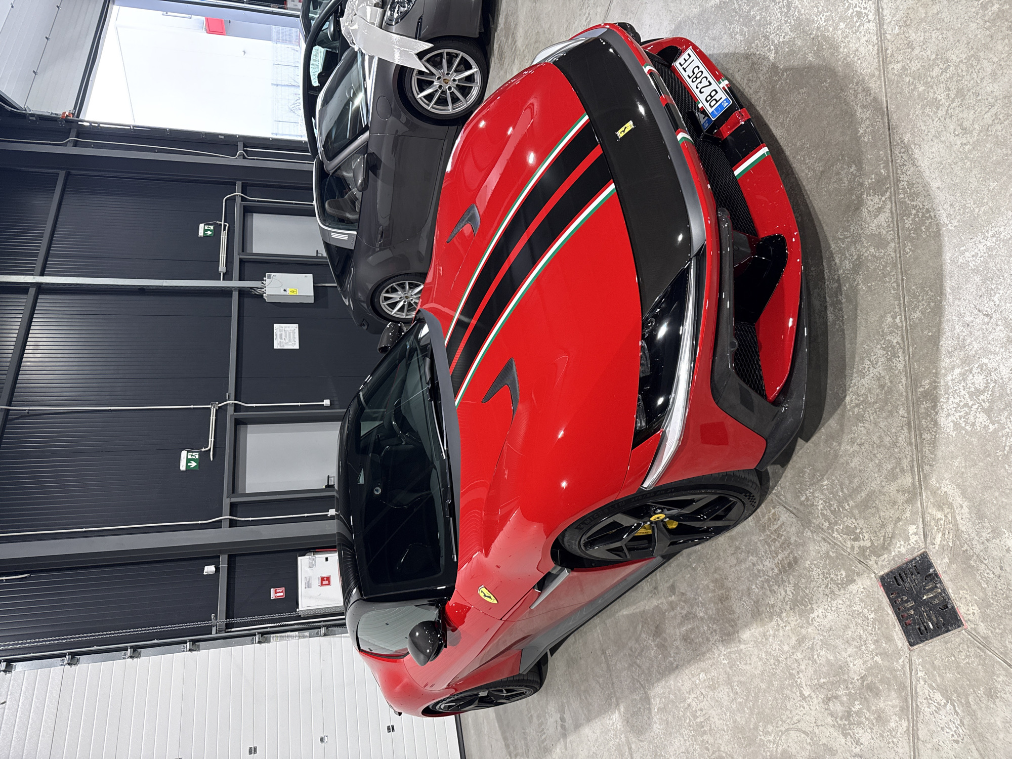 The image shows a red Ferrari with a black roof and black and green racing stripes on the hood, parked indoors on a concrete floor. The car is positioned in the foreground, angled towards the left. In the background, there is a gray Porsche Targa with a large white bow on its hood, suggesting it might be a gift or a display. The environment appears to be a car dealership or showroom, with industrial-style walls and large garage doors. The license plate of the Ferrari is visible and reads "PB 2385 TE". The scene is well-lit, highlighting the vehicles. There are no people visible in the image. The location of Plovdiv, Bulgaria, is not explicitly indicated in the image itself, but if we assume the context provided in the prompt, the scene is situated within that city.