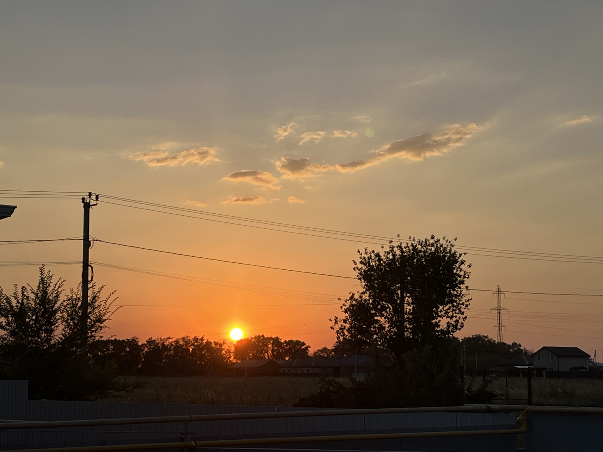The image depicts a sunset in Taganrog City District, Russia. The sun, a bright orange orb, is low on the horizon, casting a warm glow across the sky. The sky is a gradient of soft oranges, yellows, and blues, with wisps of clouds scattered across it. In the foreground, silhouetted against the vibrant sky, are trees, power lines, and a distant building. The scene appears to be rural or semi-rural, with a sense of peace and tranquility.