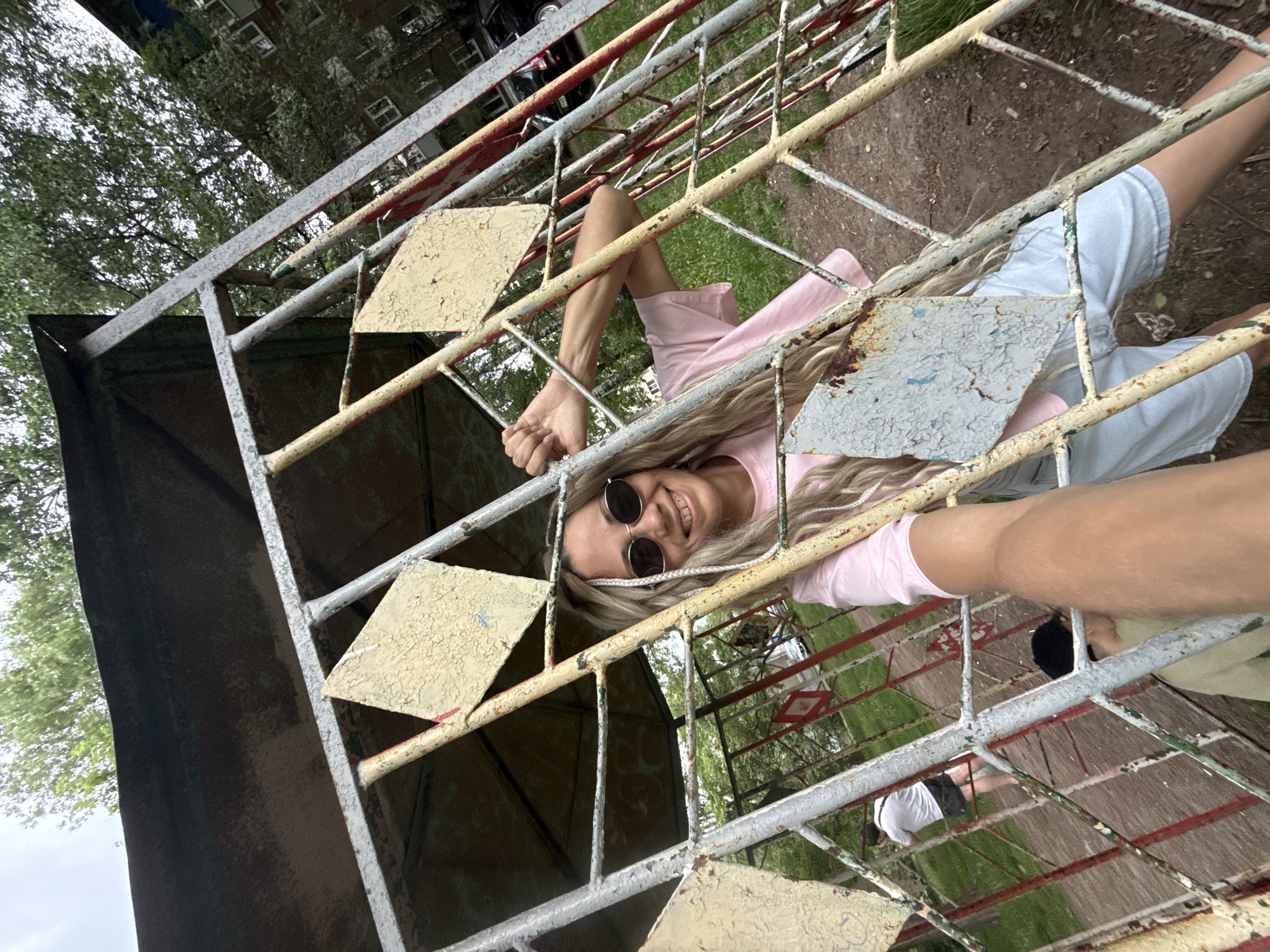 A woman with long blonde hair and sunglasses smiles at the camera. She is partially obscured by a decaying metal playground structure painted white and yellow. She wears a pink top and light-colored shorts. Through the grid of the playground structure, a grassy area is visible, where two people can be seen in the distance. The location appears to be in the city of Rusanovo, Russia.