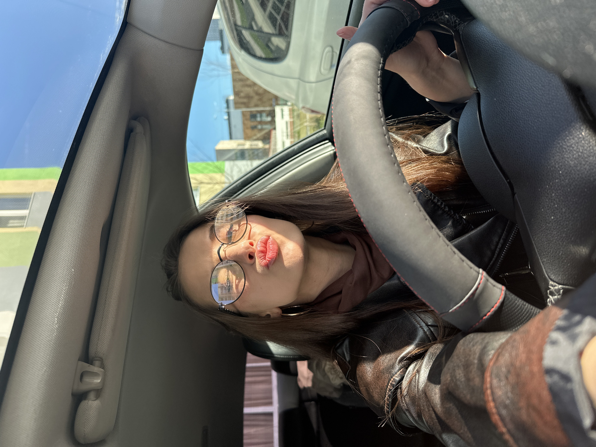 A woman is sitting in the driver's seat of a car. She is wearing a brown leather jacket, a brown turtleneck, and round-frame glasses. She is looking at the camera with pursed lips, holding the steering wheel with her left hand. The interior of the car is visible, including the grey dashboard and the sun visor. Through the car window, buildings are visible, suggesting an urban setting. The reflection in her glasses indicates buildings that possibly place her in Yekaterinburg, Russia.
