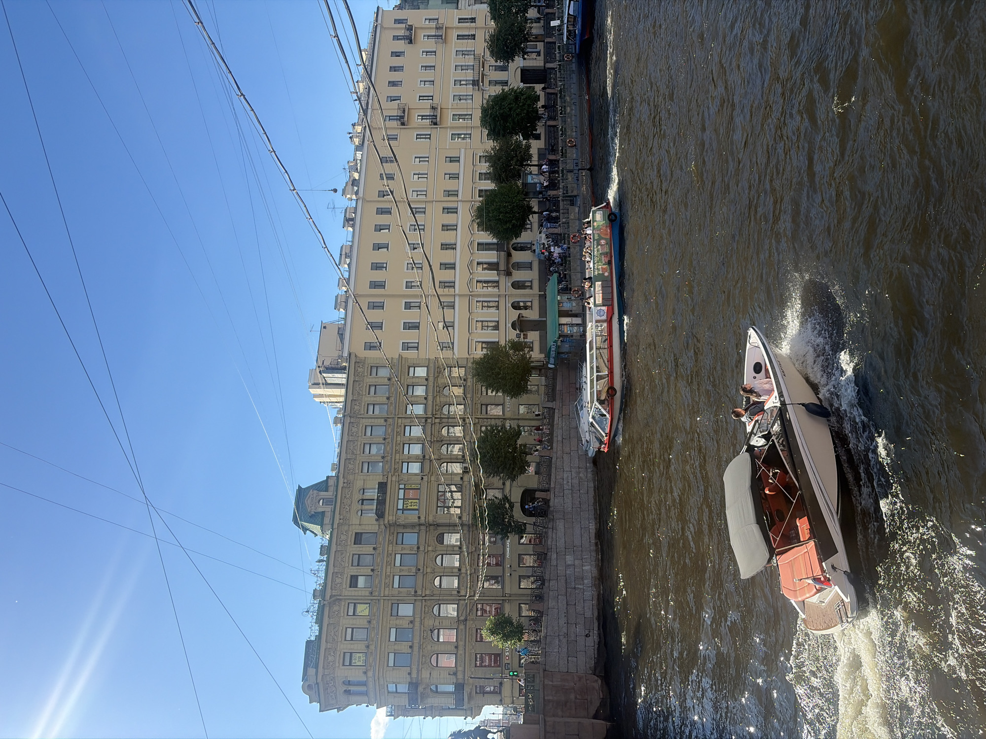 This image depicts a sunny day along a canal in Saint Petersburg, Russia. In the foreground, a white motorboat with a black canopy speeds through the dark, choppy water, leaving a white wake. Two people are seated inside the boat. Further in the background, a larger tour boat with a red and blue hull is also moving along the canal, carrying several passengers. Both sides of the canal are lined with ornate, multi-story buildings, typical of Saint Petersburg architecture, with light yellow facades and numerous windows. Trees are planted along the canal bank, providing some greenery. Overhead, a complex network of power or telephone lines crisscrosses the bright blue sky. The sunlight creates shimmering reflections on the water's surface.