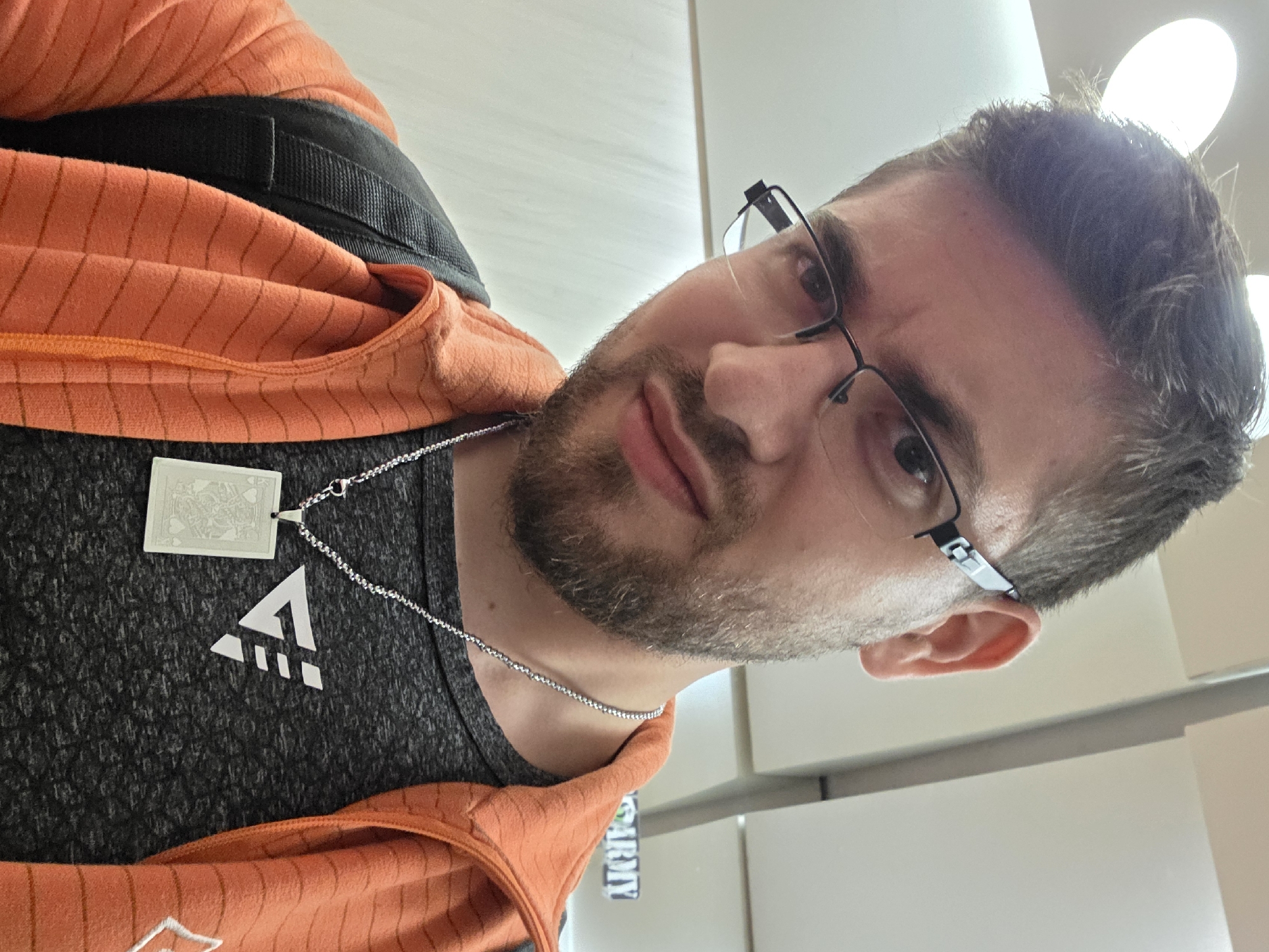 A close-up, tilted selfie depicts a light-skinned male with short brown hair and a trimmed beard. He wears black-framed glasses and is dressed in a dark grey t-shirt with a white logo consisting of an upward-pointing triangle over three dots. An orange jacket is partially visible over the t-shirt. He is wearing a silver chain necklace with a rectangular playing card pendant. A black strap is visible over his left shoulder. The background appears to be an indoor setting with white walls. A hanging sign with the text "ARMY" is partially visible in the background. There is no indication of a specific location in Plovdiv, Bulgaria.