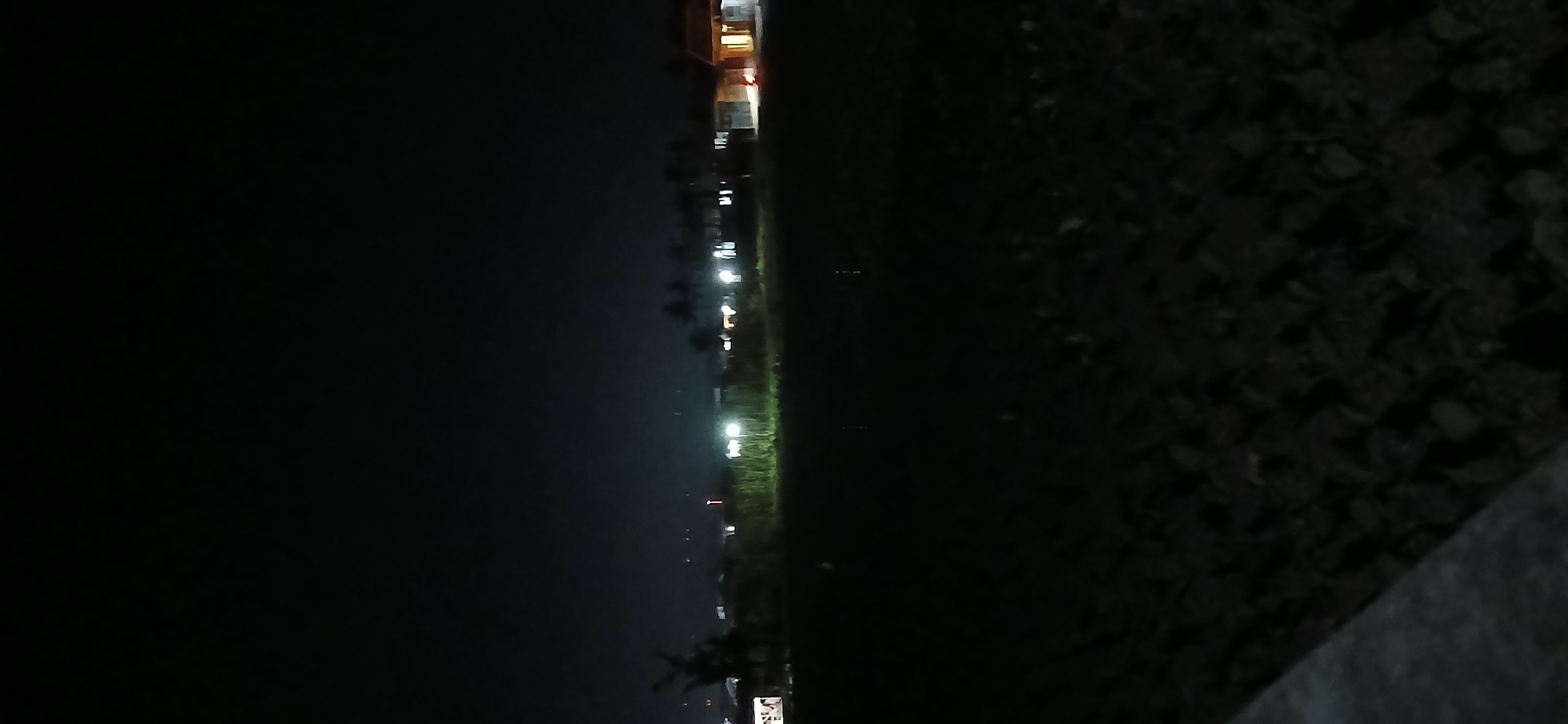 This is a nighttime photograph taken in Sekarputih, Indonesia. The scene depicts an open, dark field in the foreground, with what appears to be tilled earth, possibly a farm or undeveloped land. In the background, a line of buildings and trees are illuminated by artificial lights. These lights suggest a residential or commercial area on the outskirts of the settlement. The sky is dark and clear, typical of a rural nighttime environment. The overall impression is one of quiet darkness broken by scattered points of light.