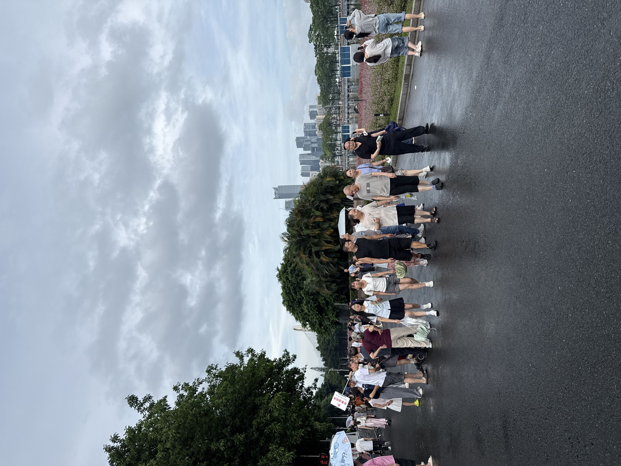 The image depicts a crowd of people walking on a wet asphalt path in Tianhe District, China, under a cloudy sky. In the background, a lake or body of water is visible, bordered by a row of trees and a cityscape with modern buildings. People are dressed casually in summer attire, with some holding umbrellas. The atmosphere appears to be that of a public gathering or stroll in a park.