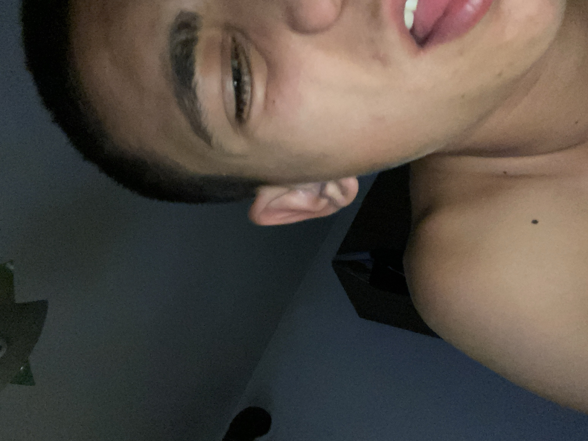 The image shows a close-up of a young person's face and shoulder, taken in low light conditions. The person is looking towards the camera with their mouth slightly open, revealing their tongue and teeth. Their right eye is partially closed. The background is dark and out of focus, with what appears to be a wall and a ceiling visible. There are no identifiable landmarks or contextual clues within the image that would suggest a specific location in Brashlen, Bulgaria. The scene is a personal snapshot rather than a depiction of a public space or event.