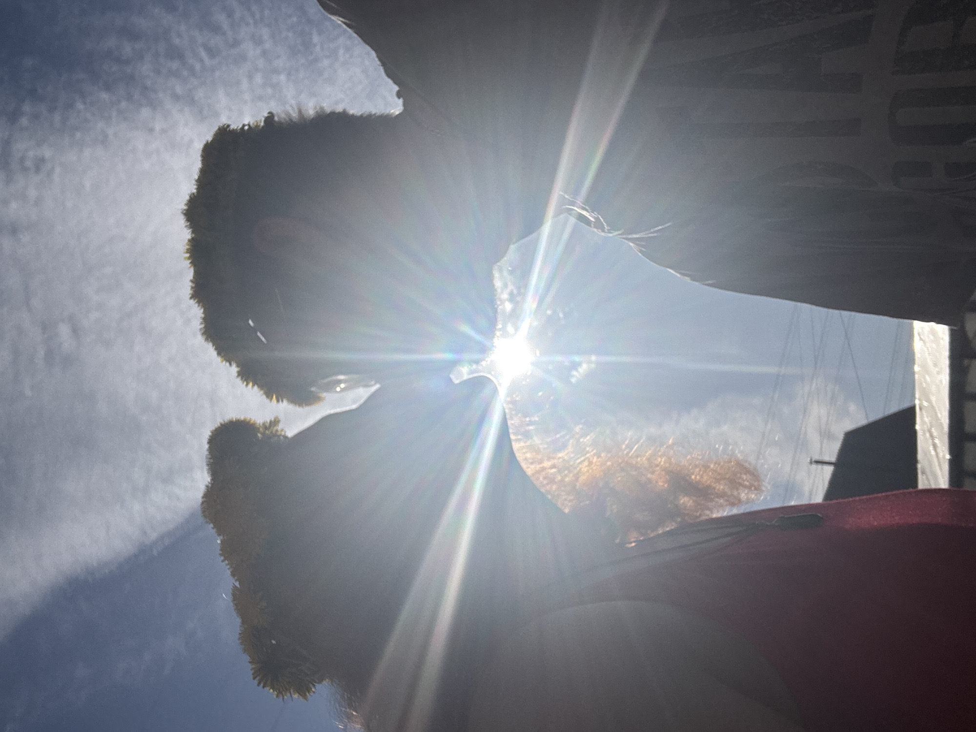 Two people are close together with the sun directly between them. The sun is very bright, causing the image to be overexposed and making it difficult to see details. The person on the left has dark curly hair. The person on the right has reddish-brown hair. A red object is visible near the bottom of the frame. There is no readily discernible location in Chaikovsky, Russia.