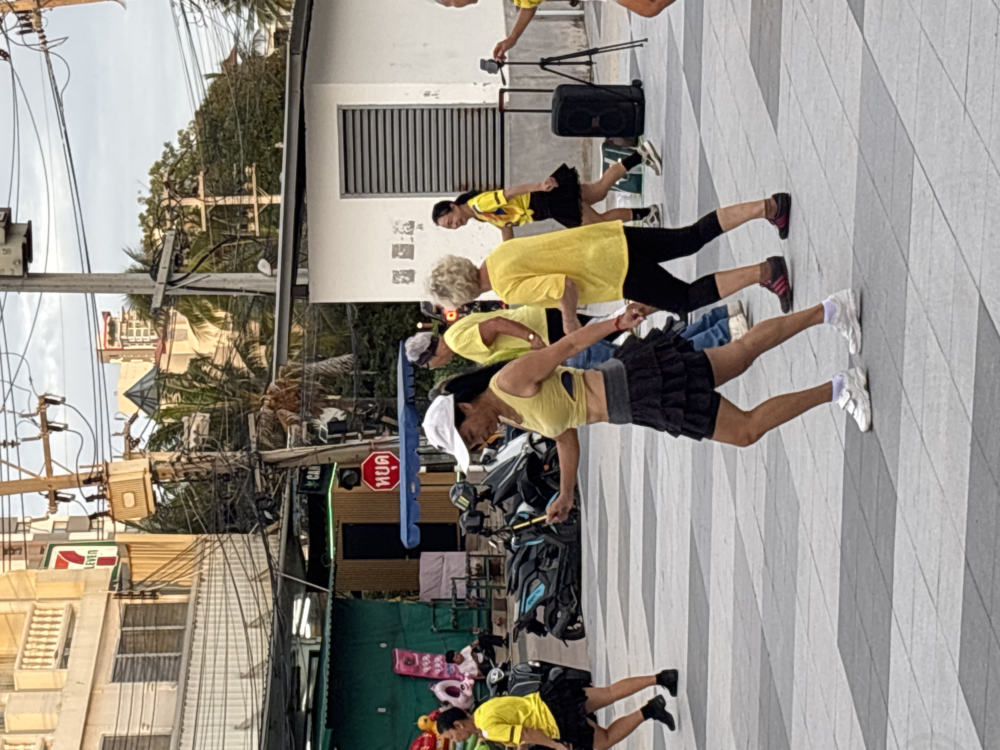 A group of women are gathered in a public square in Pattaya City, Thailand, participating in what appears to be a synchronized exercise or dance session. The women are predominantly dressed in yellow tops and black skirts or pants. One woman in the foreground, wearing a white cap and a yellow tank top, is holding a mobile device, possibly for music or recording. Behind her, other women in similar attire are engaged in the activity. The environment includes a paved square with a distinctive checkered pattern, some parked motorcycles, and buildings in the background, including a Seven-Eleven convenience store. Electrical wires and utility poles are visible overhead, typical of urban streetscapes in Thailand. The overall scene suggests a casual, outdoor recreational activity taking place in an urban setting.