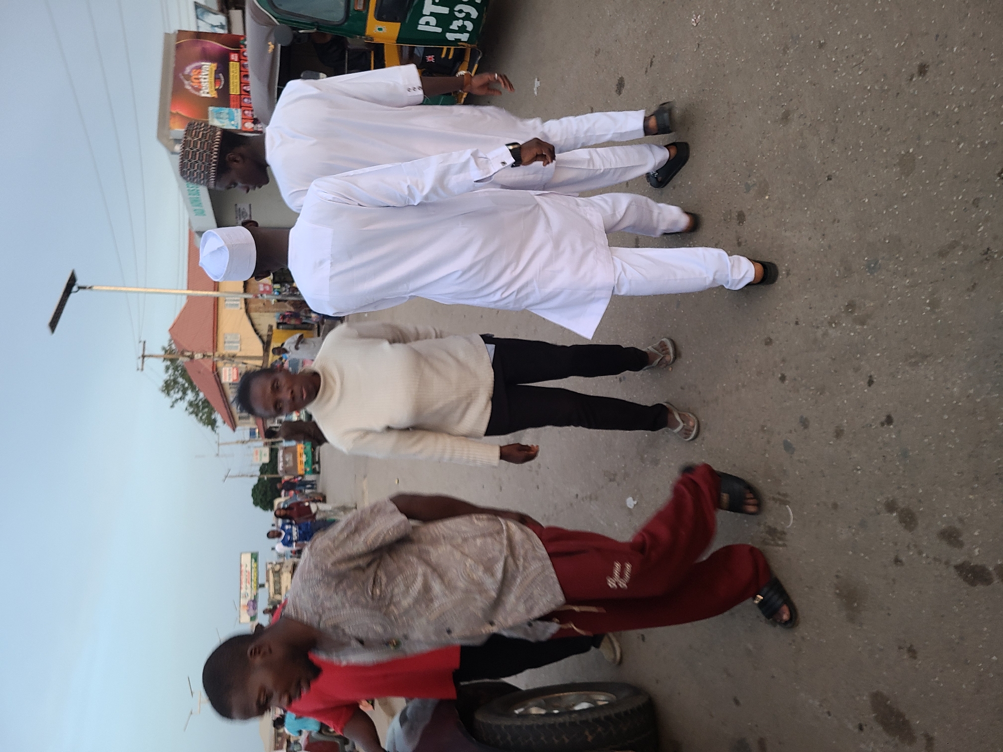The image shows an outdoor scene in an unidentified city. Two men in white traditional attire stand on the right. To their left, is a person in a white sweater and black pants. Closer to the foreground, a person in a gray shirt and red pants is to the left and a person in a red shirt is on the bottom left with a tire in front of them. In the background, there are buildings, utility poles, and other people.