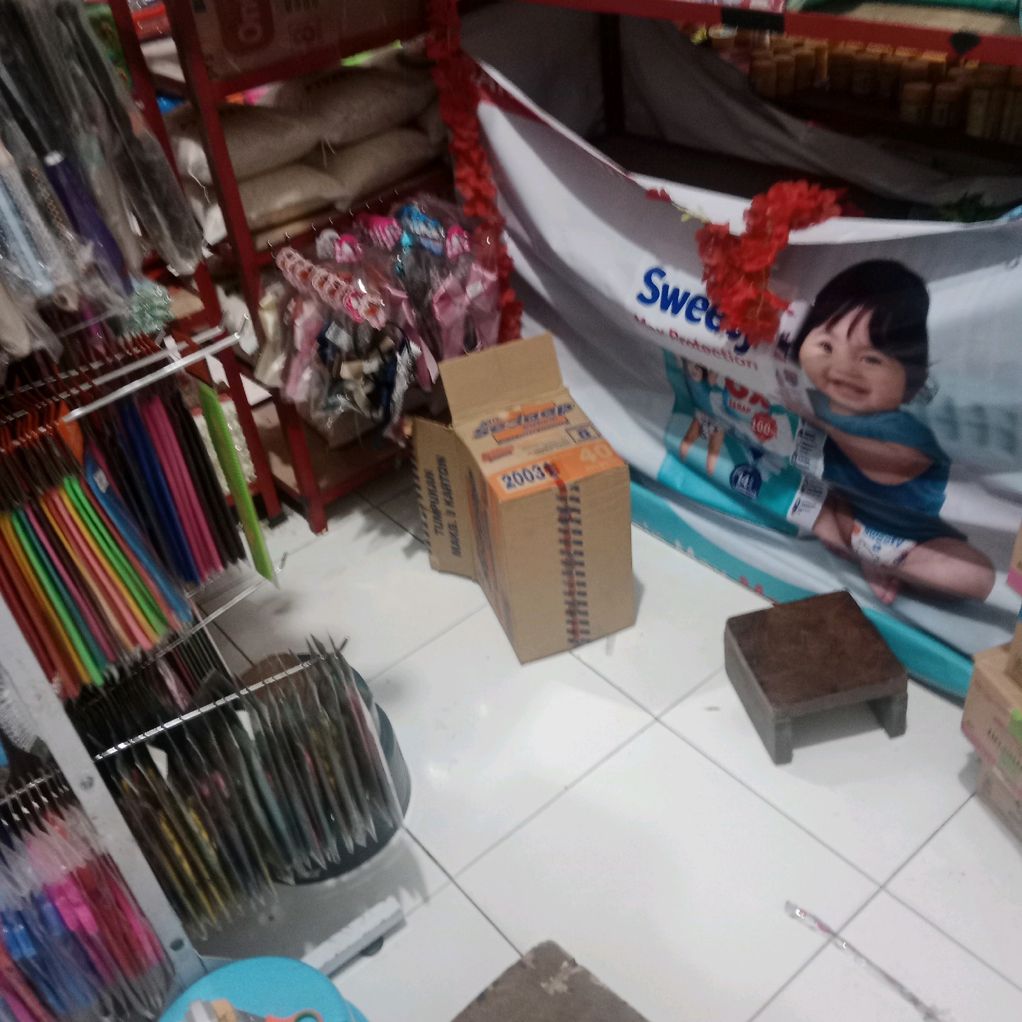 The image depicts an indoor retail environment, likely a small shop or market stall, exhibiting a variety of merchandise. To the left, an assortment of items is displayed on metal racks, including what appear to be colorful bundles of fabric or streamers, possibly for decoration or crafts, and other packaged goods. A red metal shelf unit supports some of these items.
In the center of the scene, a cardboard box with orange and white labeling is placed on the floor. The box has text and numerical indicators such as "2003" and "40" visible. Behind this box, a large white banner featuring an image of a smiling baby and the word "Swee..." is prominently displayed, adorned with red decorative flowers.
To the right, a small, dark brown wooden stool or step is situated on the tiled floor. Further to the right, additional packaged items are visible on shelves, and a black fabric covers a portion of the background. The floor is tiled with light-colored squares, and the overall lighting suggests an indoor setting.
This scene is located in Tegowanu Wetan, Indonesia, a specific location within the country.
