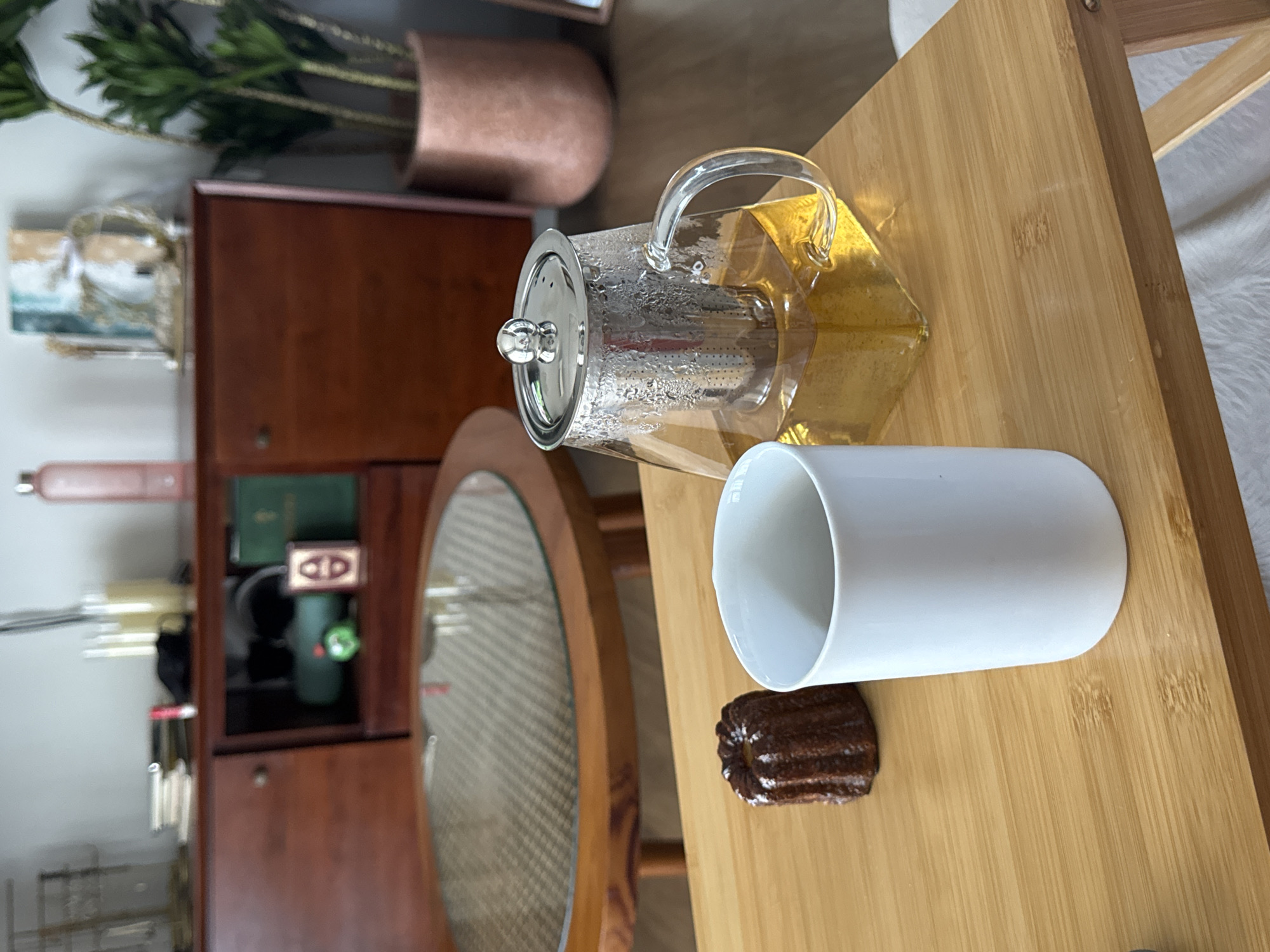 A close-up, high-angle shot captures a bamboo table with a canelé pastry and a white mug. To the right, a glass teapot filled with amber liquid sits, its metal infuser visible. In the background, a wooden cabinet with a glass-top coffee table is visible, as well as a potted plant and various decorative items. The scene appears to be indoors, possibly in a cafe or residential setting in North Kuta, Indonesia.