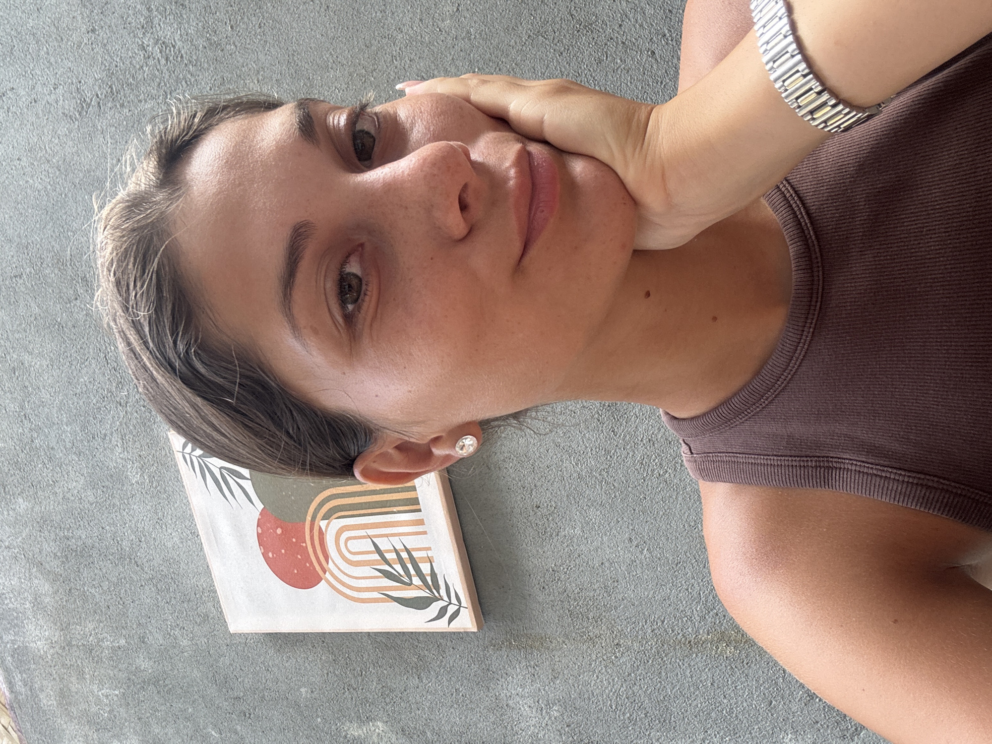 A woman is holding her face in her hand. She is wearing a brown tank top and a silver watch. There is a framed picture with an abstract design on it next to her head. The background is a gray concrete surface. It is not possible to determine the location as Đà Nẵng, Vietnam, from the given information.