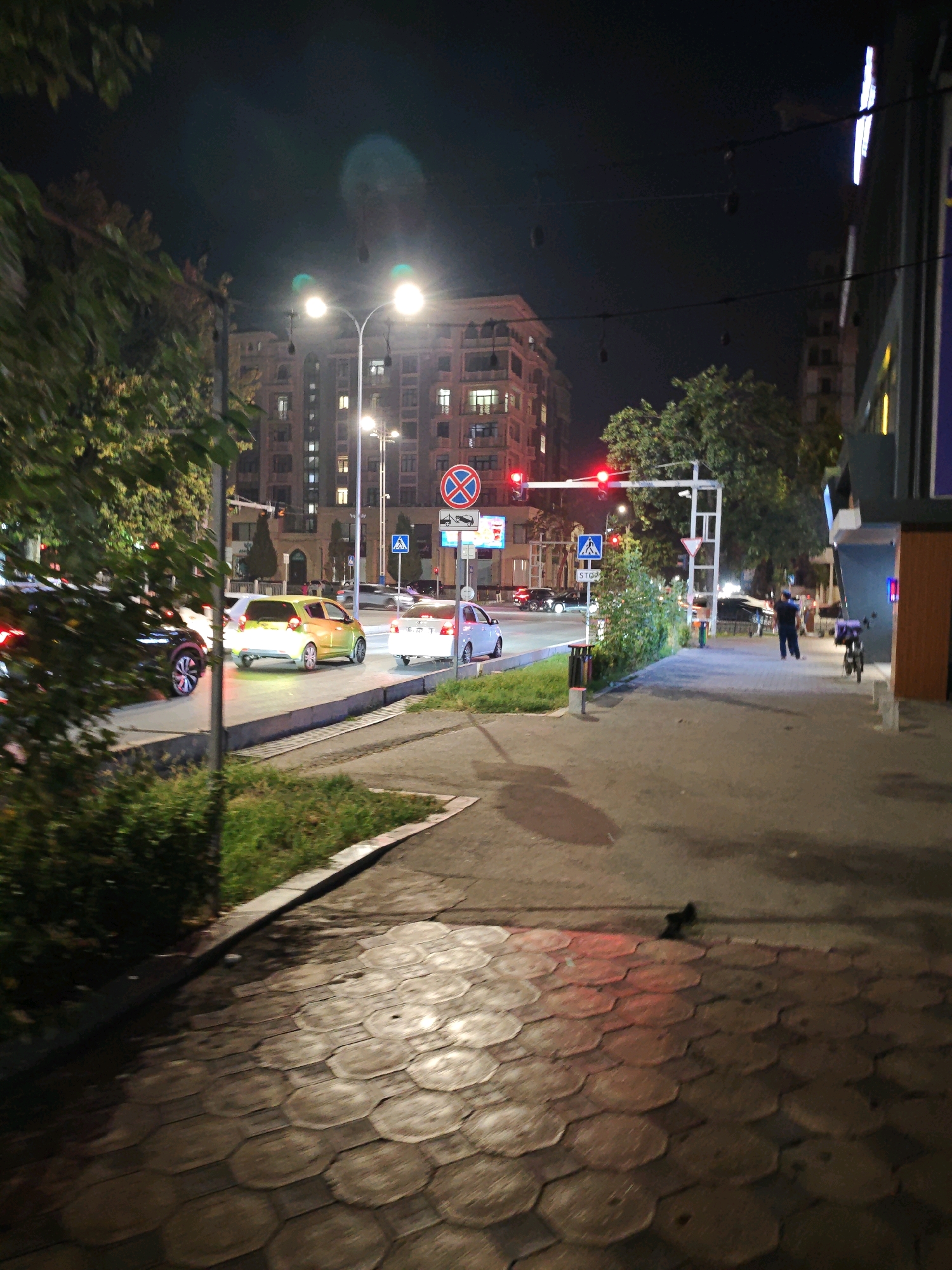 The image captures a street scene at night in Tashkent, Uzbekistan. A multi-lane road is visible with several cars, including a yellow hatchback in the foreground. Traffic lights are red, indicating a stop. Pedestrian crossings are marked by blue signs. The sidewalk is paved with hexagonal tiles. Buildings line the street, some with illuminated windows, suggesting residential or commercial spaces. Trees and bushes add greenery to the urban landscape. A few people are present, one of whom is walking on the sidewalk, and a motorcycle is parked nearby. Streetlights illuminate the area. The overall atmosphere is that of a busy urban environment at night.