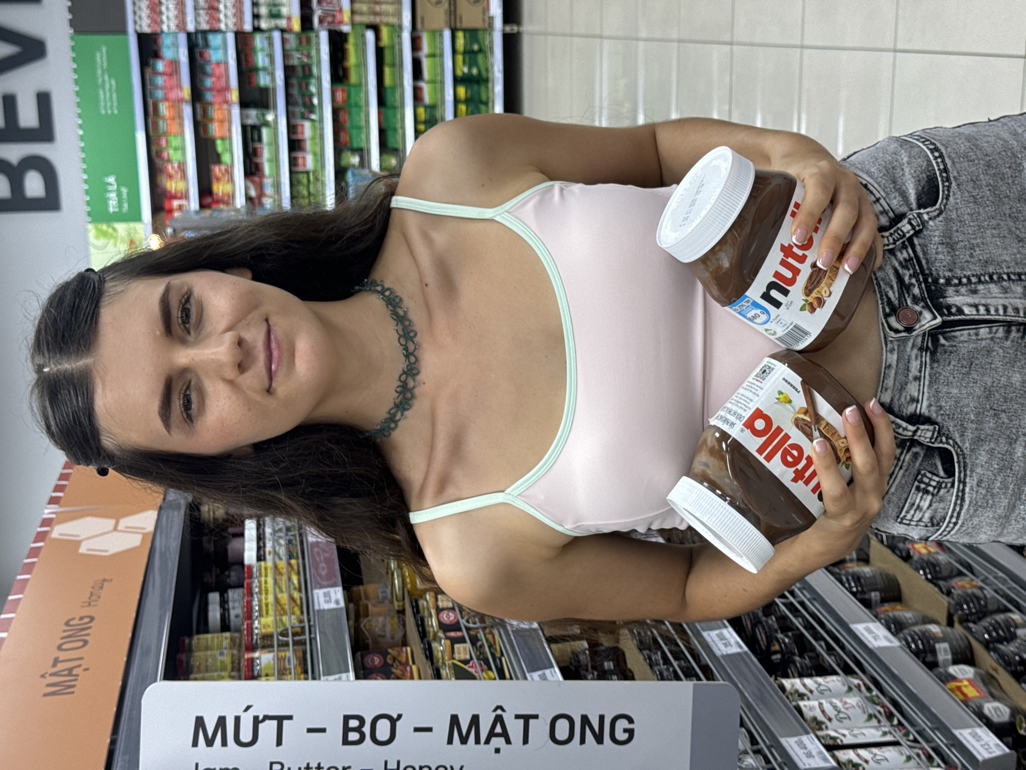 The image depicts a woman holding two jars of Nutella in a store aisle. The aisle appears to be in the city of Phu Thuy Ward, Vietnam, based on the Vietnamese signage visible, which reads "MỨT – BƠ – MẬT ONG Jam - Butter - Honey" and "MẬT ONG Horany," and "TRÀ LÀ" on a green sign. The woman, who is looking at the camera with a slight smile, is wearing a light pink tank top with light green trim and grey acid-wash jeans. She is holding a jar of Nutella in each hand, with the labels visible. The shelves behind her are stocked with various food items, and there are rows of Nutella jars on the shelves to her right. The overall lighting in the store is bright.