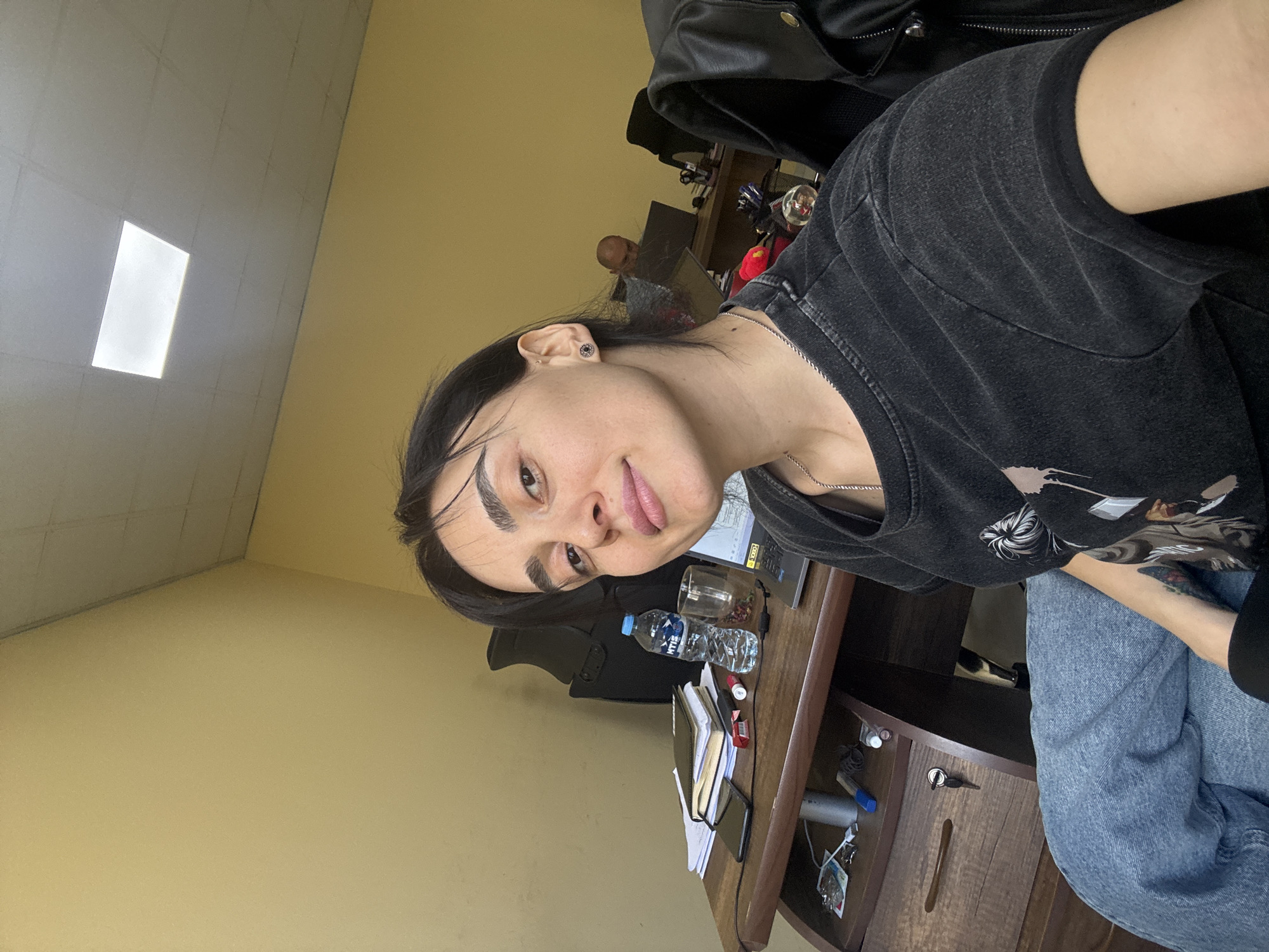 A medium indoor shot shows a light-skinned individual with dark hair and eyebrows. The person is wearing a dark graphic t-shirt and light blue jeans. A black leather jacket hangs on a chair behind. The background is a yellow painted wall with a rectangular light. The person is at a dark wooden desk with a water bottle, paperwork, a book and a laptop. The location appears to be indoors in Tbilisi, Georgia.