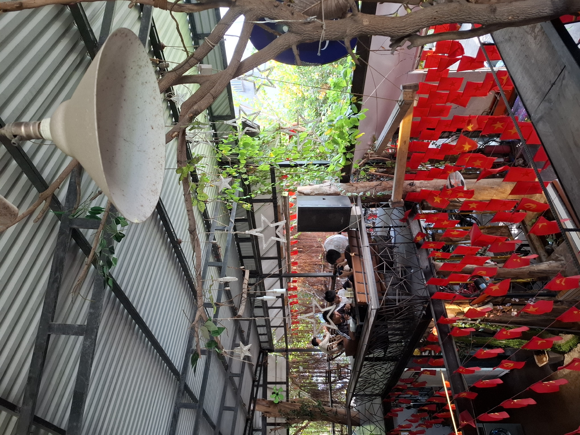 The image shows an indoor/outdoor seating area of a cafe in Nha Trang, Vietnam, possibly a rooftop or elevated level. The structure has a corrugated metal roof supported by dark metal beams. The space is decorated with numerous small red flags with yellow stars, strung across railings and hanging from overhead supports. Greenery, including vines and tree branches, is integrated into the decor, with a large tree trunk visible in the foreground on the right. A large, conical industrial-style lamp hangs from the ceiling. In the background, several people are seated at tables, some appearing to be dining or socializing. One group of young men is looking at phones. The overall atmosphere appears casual and somewhat rustic, with a blend of natural elements and simple construction.