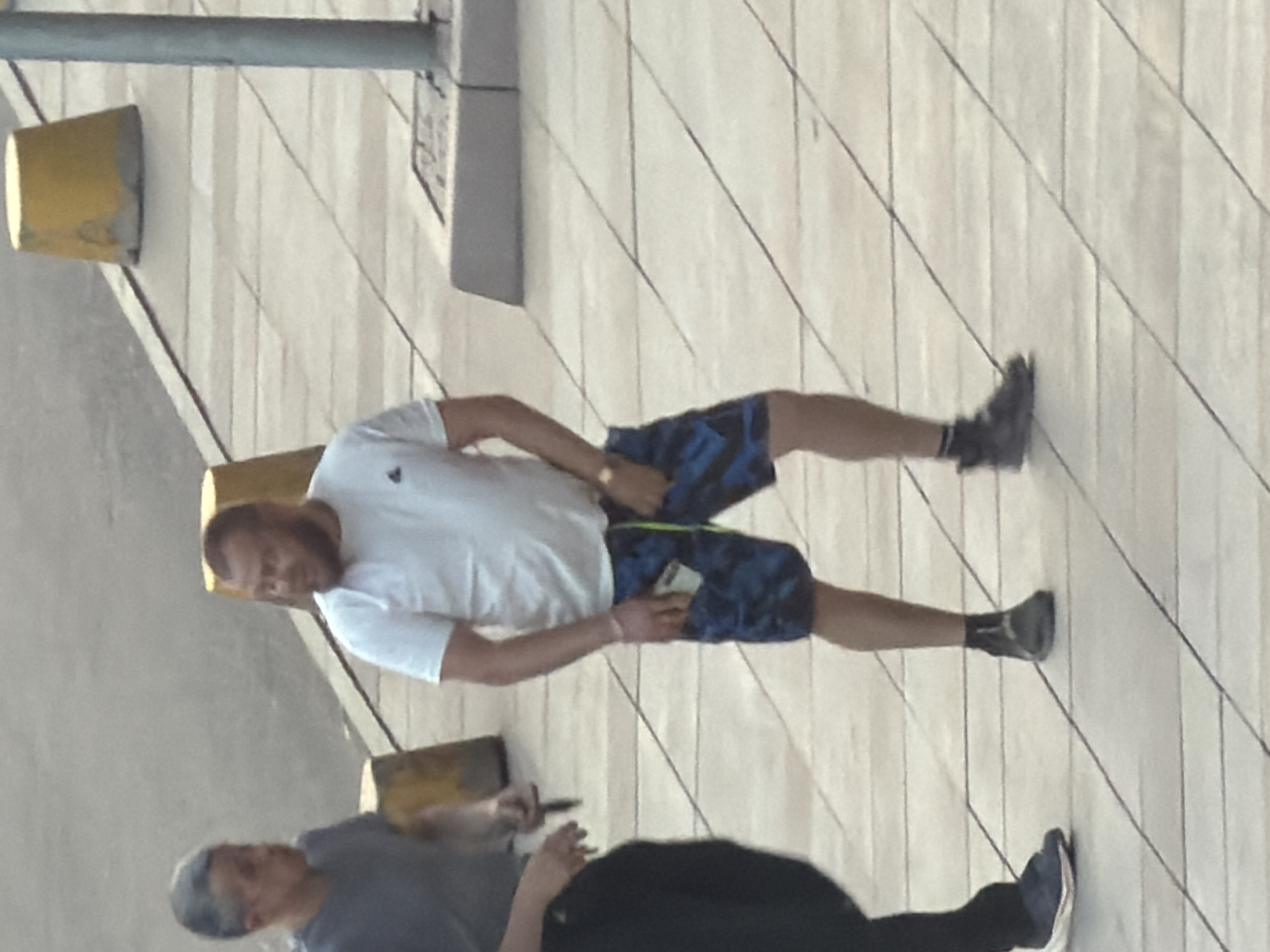 Two men are walking on a tiled sidewalk in Krasnoyarsk, Russia. The man on the left is wearing a dark t-shirt and dark pants, and the man on the right is wearing a white t-shirt and blue patterned shorts. The man on the right is holding a phone in his hand. There are yellow traffic cones visible in the background.