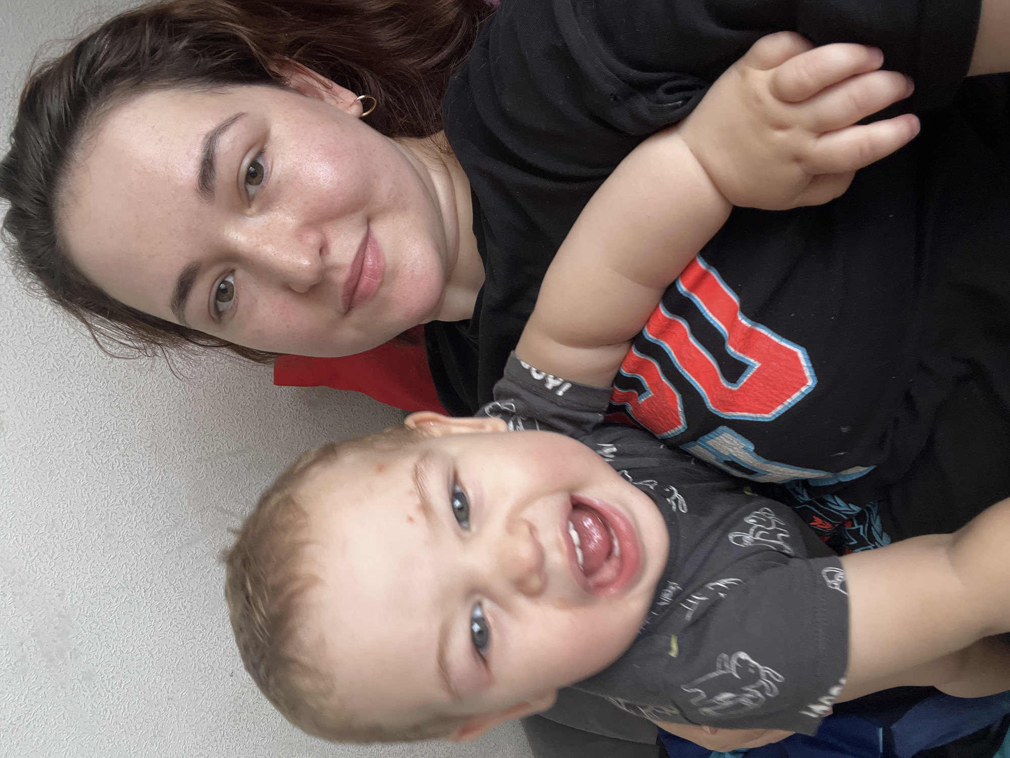 A woman with brown hair and a black t-shirt is holding a baby. The baby is wearing a grey t-shirt with white animal graphics. The woman is smiling at the camera, and the baby is looking away with its mouth open. The background is a textured white wall. The image is taken from a close-up perspective. No specific location within Chelyabinsk, Russia, can be determined from the visual information provided.