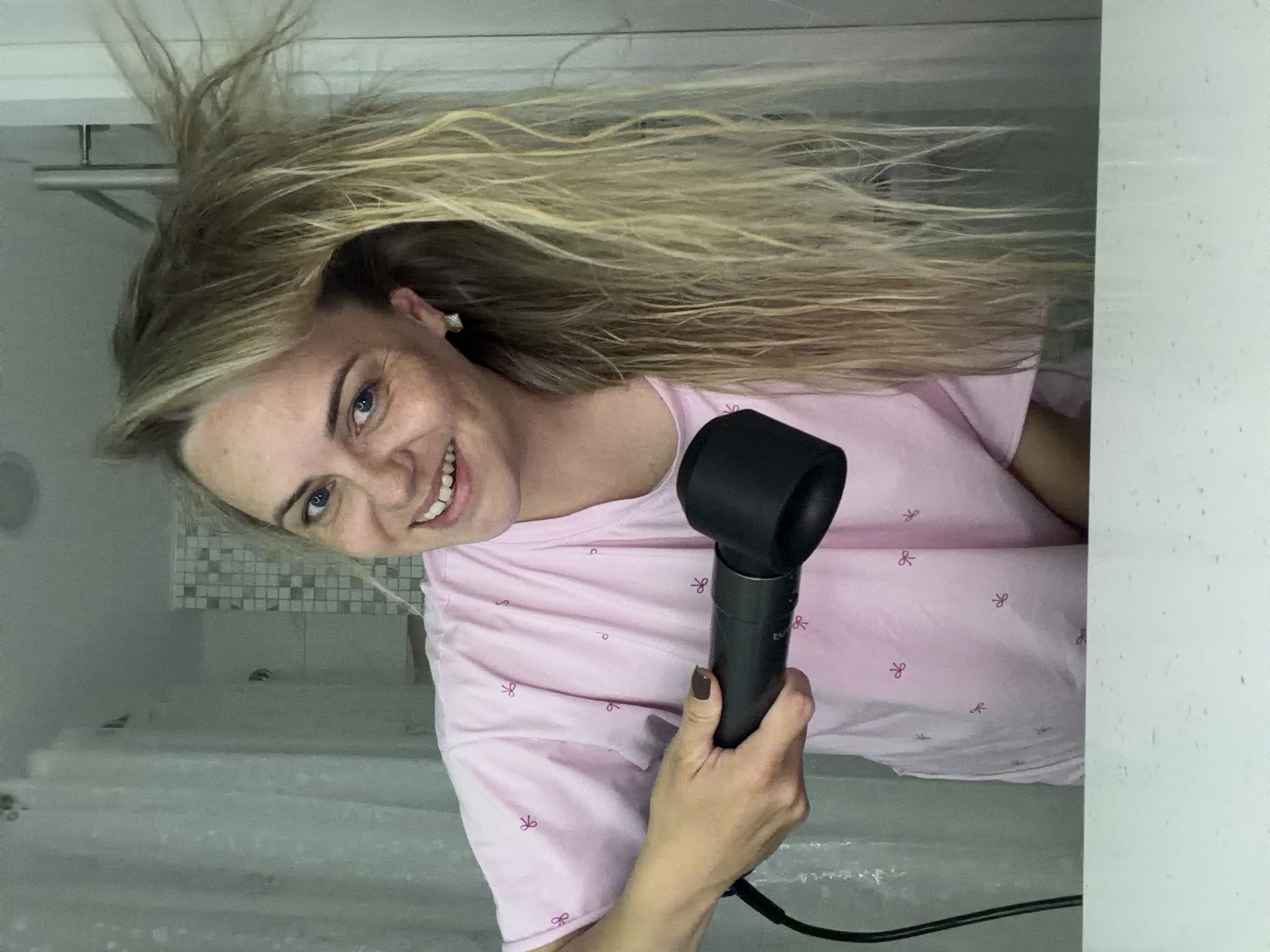 An adult Caucasian female is in a bathroom, likely her own, and is using a hair dryer to dry her wet, blonde hair. She is wearing a pink t-shirt with a subtle bow pattern. Her hair is long and appears to be somewhat messy from being wet. She is smiling at the camera. The background shows a shower curtain on the left and tiled walls on the right, indicative of a bathroom environment. The scene is lit by artificial lighting. There is no visible indication that this image was taken in Votkinsk, Russia, or any specific city.