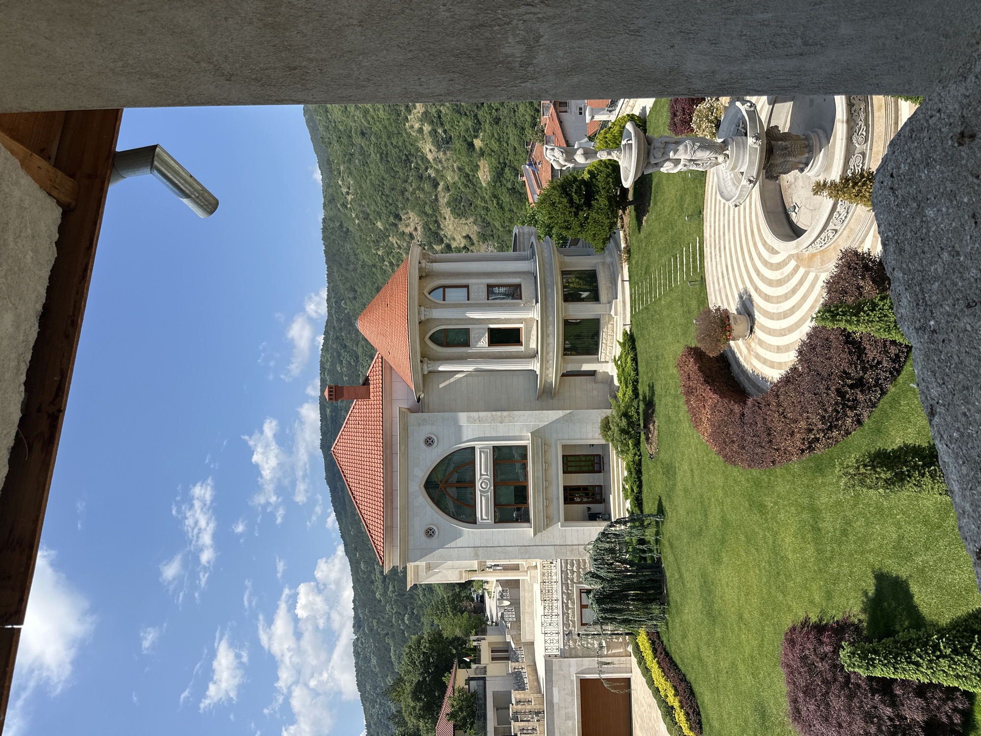 The image displays a large, elaborate residential property with an expansive garden, situated against a backdrop of forested mountains under a clear blue sky with scattered white cumulus clouds. The scene is illuminated by direct sunlight, casting short shadows indicative of daytime. The location is Asenovgrad, Bulgaria. In the immediate foreground, a rough, textured concrete or stone wall is visible on the right, extending vertically, framing the view. A flat, rough stone surface also occupies the bottom central and right edge. Above, a wooden beam and a white, possibly insulated, ceiling structure are visible. A silver metal rain gutter outlet pipe projects downwards from the upper right corner. The central subject is a multi-story, light-colored (cream or pale beige) house with a prominent reddish-orange tiled roof. The main section features a large, arched window on the upper level, flanked by two small, round decorative vents. Below this, the ground floor has a recessed entrance with two dark-paneled doors and a few steps leading to the lawn. To the left, a lower section of the house includes a wooden garage door and paved driveway, alongside terraces with dark metal railings. To the right of the main section is a distinctive round tower structure with a conical reddish-orange roof. This tower features multiple arched windows on at least two levels, interspersed with decorative columns. The extensive garden surrounding the house is meticulously maintained, featuring a vibrant green lawn. Various decorative shrubbery and plants are arranged throughout. A long bed of yellow and green shrubs runs alongside the driveway on the left. A large, weeping-form tree with green foliage stands directly in front of the main house section. Further to the right, a curved bed of dark reddish-brown shrubs borders a circular paved area. This paved area displays concentric patterns of alternating light and dark stripes. Within this circular area stands a multi-tiered stone fountain, adorned with figural sculptures, including female figures supporting an upper basin, and decorative relief work on the lower basin. The fountain appears inactive. A series of light-colored, rectangular stepping stones forms a path across the lawn. In the mid-ground and background, dense green forests cover rolling mountains, indicative of a hilly or mountainous terrain. Other residential buildings with reddish-orange tiled roofs are visible amidst the trees in the distance, particularly to the right of the main house, suggesting a populated area within this natural setting. No individuals are