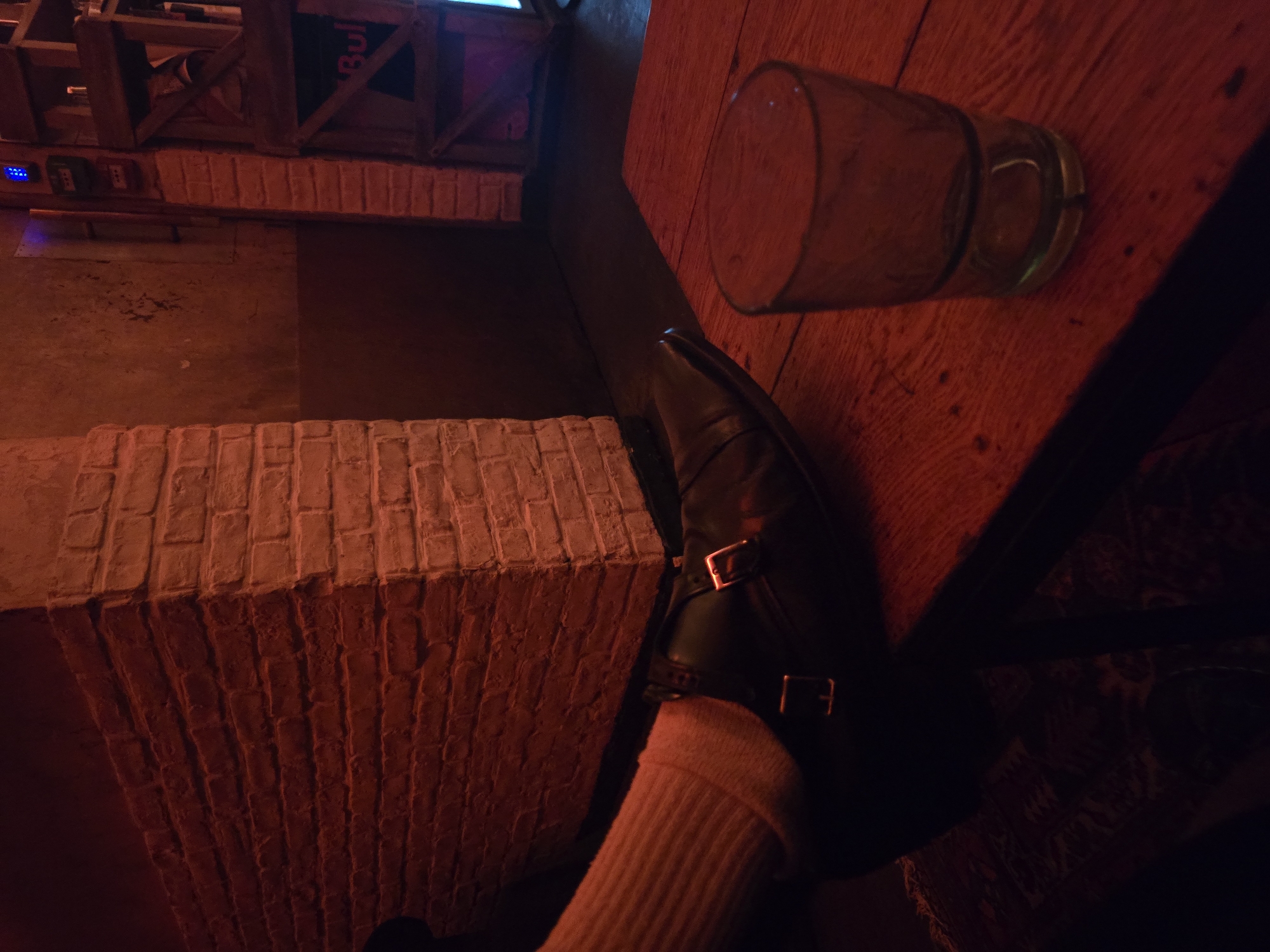 The image depicts a scene inside what appears to be a bar or pub, possibly located in Paris, France, given the architectural features. A brick-like structure, possibly a wall or a part of the interior design, is visible. A leg clad in a pink sock and a black leather shoe with buckles is resting on the structure. Nearby, a glass is lying on a wooden table. Above, there's an object that looks like a wooden crate. The lighting is dim and warm, giving the scene a cozy and intimate feel.