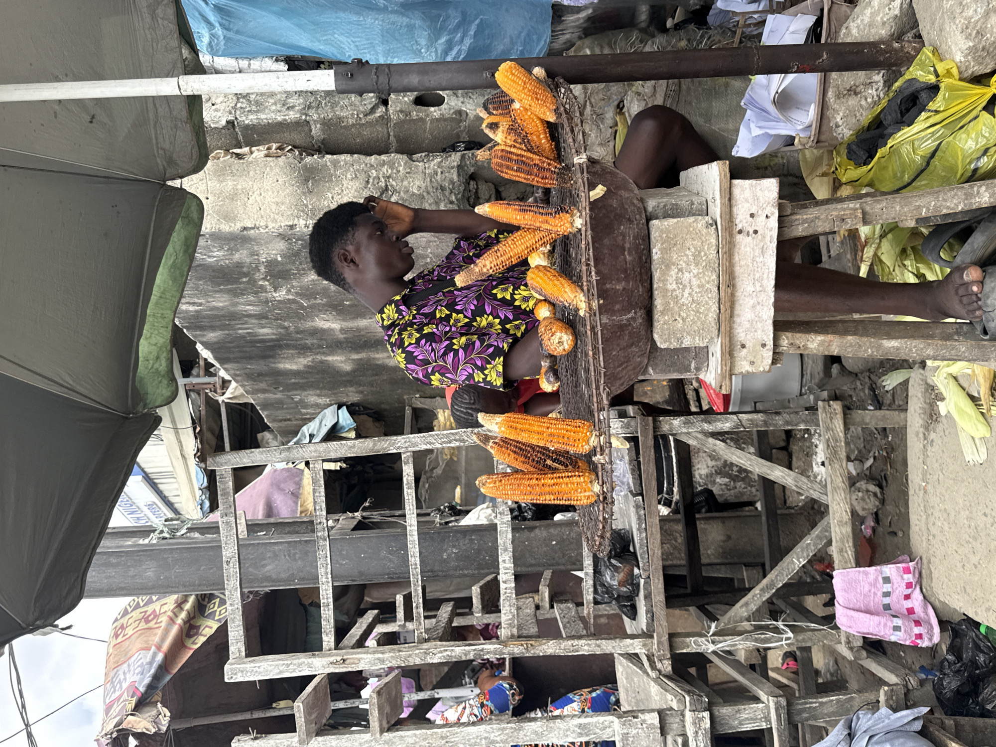 In Lekki, Nigeria, a young Black male is positioned at a street-side vendor stall, selling roasted corn. He wears a vibrant, short-sleeved shirt with a purple, yellow, and black floral pattern. His right hand is raised to his forehead, and he appears to be looking down, perhaps in contemplation or due to fatigue.
In front of him, a metal grate is set into a large, dark bowl, which rests on a wooden platform constructed from two concrete blocks and a plank of wood. The grate holds numerous ears of roasted corn, some partially charred, displaying various stages of doneness. A few corn cobs are also visible on the ground around the stall.
The stall is made of rough, weathered wood, with a multi-tiered structure to its left, possibly for storage or displaying other goods. Colorful fabrics and tarps are draped over parts of the structure, suggesting it's an open-air market. The background consists of a rough, textured concrete wall and a large, dark umbrella providing shade over the vendor and his goods. Debris, including plastic bags, discarded materials, and some yellowing corn husks, are scattered around the base of the stall, indicating a busy, functional marketplace environment. The ground is concrete, showing signs of wear and tear.