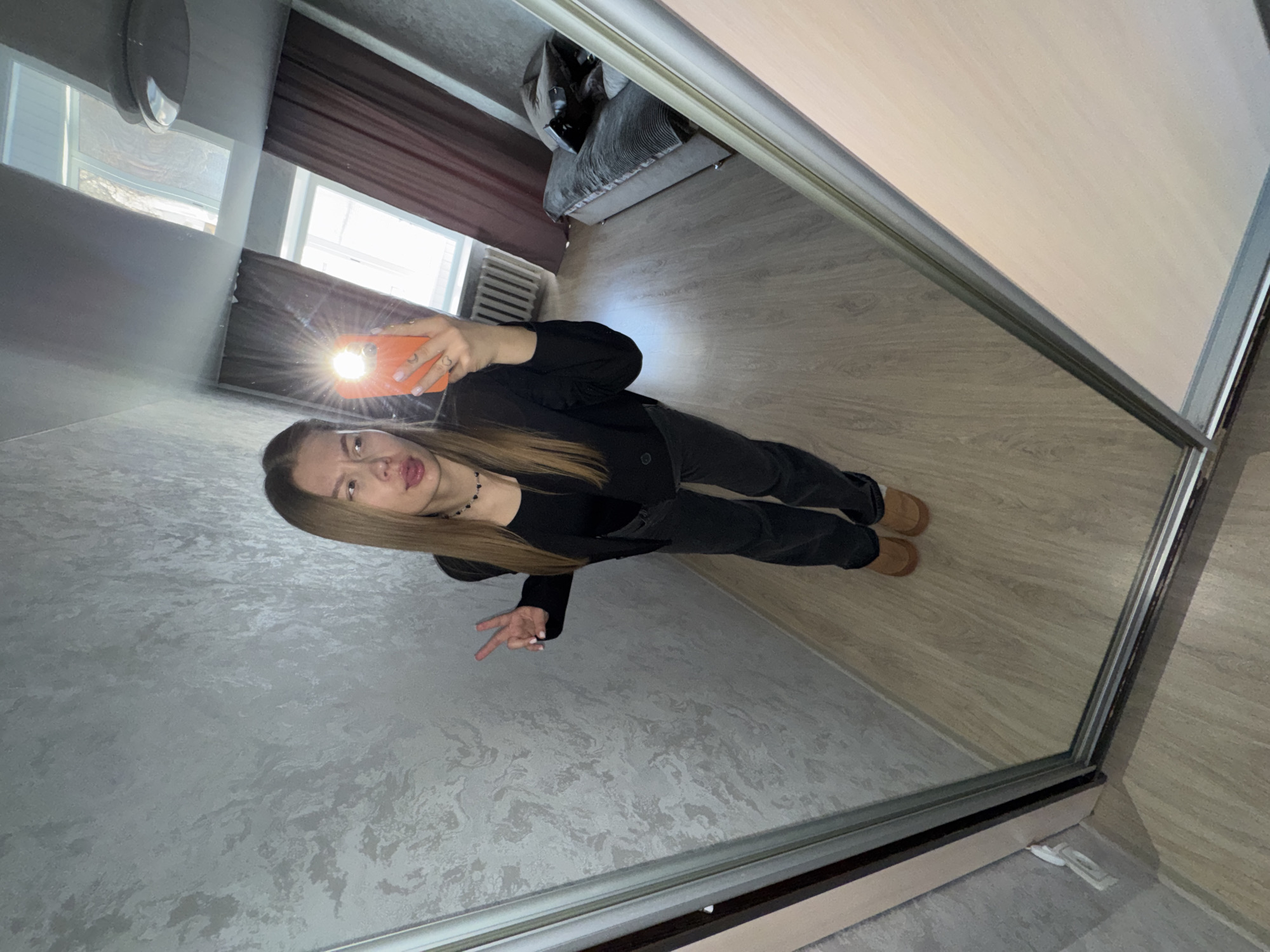 A young woman is captured taking a mirror selfie indoors. The image, which appears rotated 90 degrees, shows her standing upright, reflected in a large, full-length mirror, likely part of a wardrobe door. She has long, straight, light brown hair and is dressed in a black long-sleeved top or jacket, black pants, and light brown slip-on shoes.
She holds an orange smartphone in her right hand, aimed at the mirror, with the camera flash creating a prominent starburst effect. Her left hand is raised, forming a peace sign gesture. Her facial expression is playful, featuring a slightly puckered mouth with her tongue peeking out, and squinted eyes, as she looks towards the camera in the reflection. She wears light pink nail polish and a black choker necklace with small heart-shaped beads.
The room's interior is reflected around her. The floor is light-colored laminate or wood. A textured gray wall is visible to her left. The ceiling is also light-colored, reflecting a round light fixture. Two windows with dark brown curtains are present in the background, allowing bright natural light to enter, indicating it is daytime. Through the windows, outdoor trees are visible. A white radiator is situated beneath one of the windows. A dark gray sofa or armchair is partially visible in the far background. Near the bottom right corner of the mirror's frame, a white power outlet and cable are visible on the floor. The setting suggests a casual, residential environment in Chaikovsky, Russia. There is no visible text in the image.