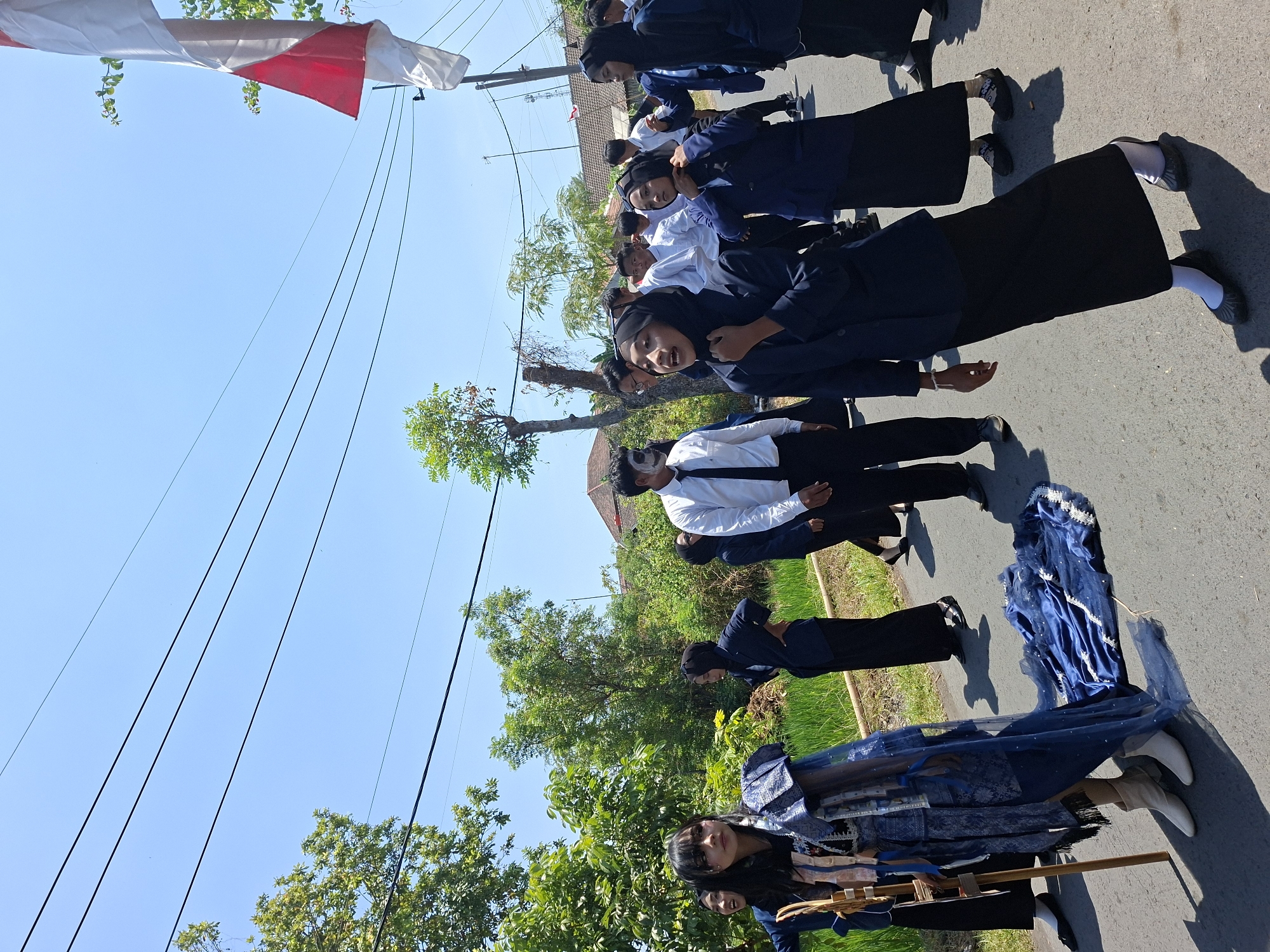 In Tongaswetan, Indonesia, a group of young people in dark blue blazers and black skirts or pants are participating in an outdoor event. One individual in the foreground, dressed in a distinctive blue and silver outfit with a long train, stands holding a wooden staff. This person has black hair and makeup resembling a mask on their face. Other individuals in the group are wearing white shirts with black ties. The background shows lush green trees and vegetation, a rice field, and houses with red tile roofs. Overhead, power lines and a partial view of the Indonesian flag on a pole are visible against a clear blue sky. The scene appears to be a procession or performance taking place on a paved road.
