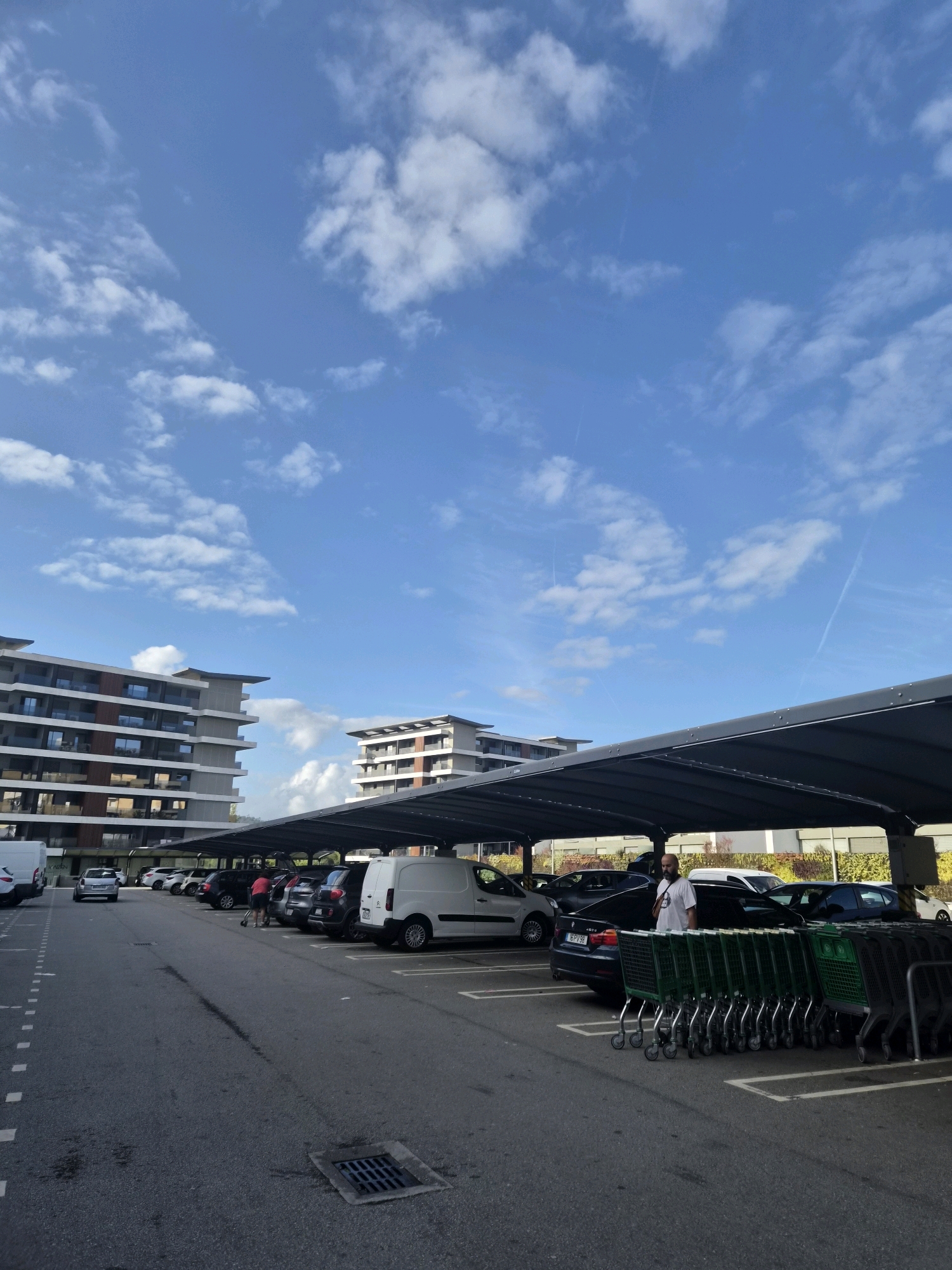 A man with a beard and a white t-shirt is standing near a line of shopping carts in a covered parking lot in Barcelos, Portugal. He is looking at a black car. To the left, there are other cars parked under the shelter. In the background, there are two modern apartment buildings. The sky is blue with scattered white clouds.
