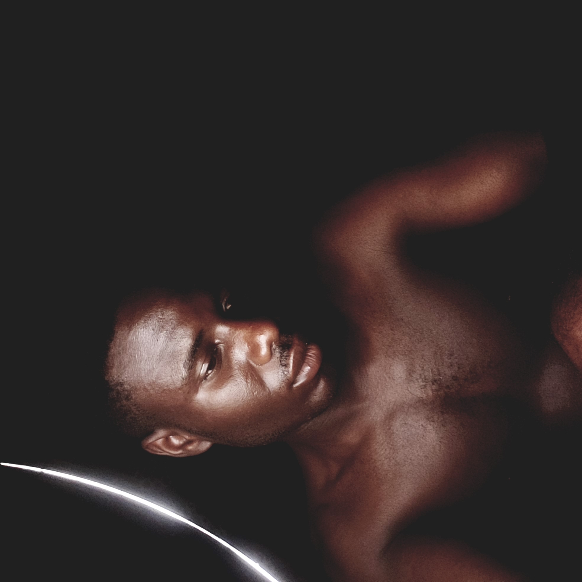 A close-up, low-key photograph depicts a shirtless dark-skinned man. He is lying on a dark surface, possibly nude, with his head turned towards the left of the frame. A curved, bright white light trails from the lower-left corner towards his face. The environment is entirely black. There is no indication of any specific location in Narayi, Nigeria.