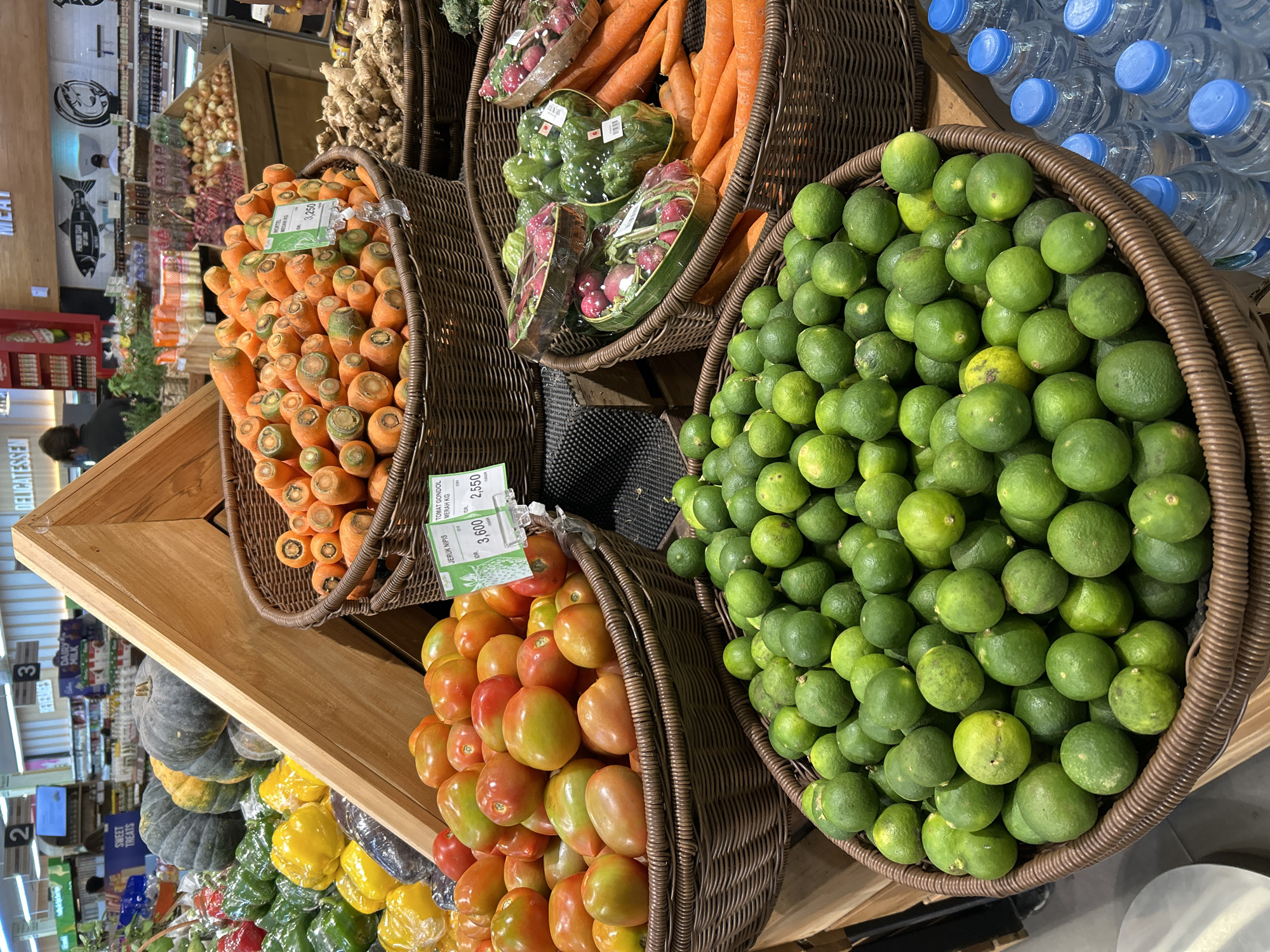 This image depicts a vibrant produce display in a supermarket, likely within the city of Ubud District, Indonesia, given the signage and the types of produce. In the foreground, a large wicker basket is overflowing with numerous small, green limes, some exhibiting slight yellowing. To the right of the limes, a collection of clear plastic water bottles with blue caps is neatly arranged. Behind the limes, another wicker basket holds a generous amount of bright red tomatoes, some with green streaks, indicating ripeness.
Adjacent to the tomatoes, a woven basket is filled with an abundance of carrots, their tops visible. To the right of the carrots, a basket contains ginger roots. Further back, another basket showcases wrapped radishes and green bell peppers. In the background, larger produce like pumpkins and gourds are displayed, alongside more carrots and other vegetables. The overall environment is a well-lit supermarket aisle, with various shelves and signage indicating product information and pricing. A few blurry figures can be seen in the distance, suggesting shoppers browsing the aisles. The display is organized and appealing, designed to attract customers with fresh produce.