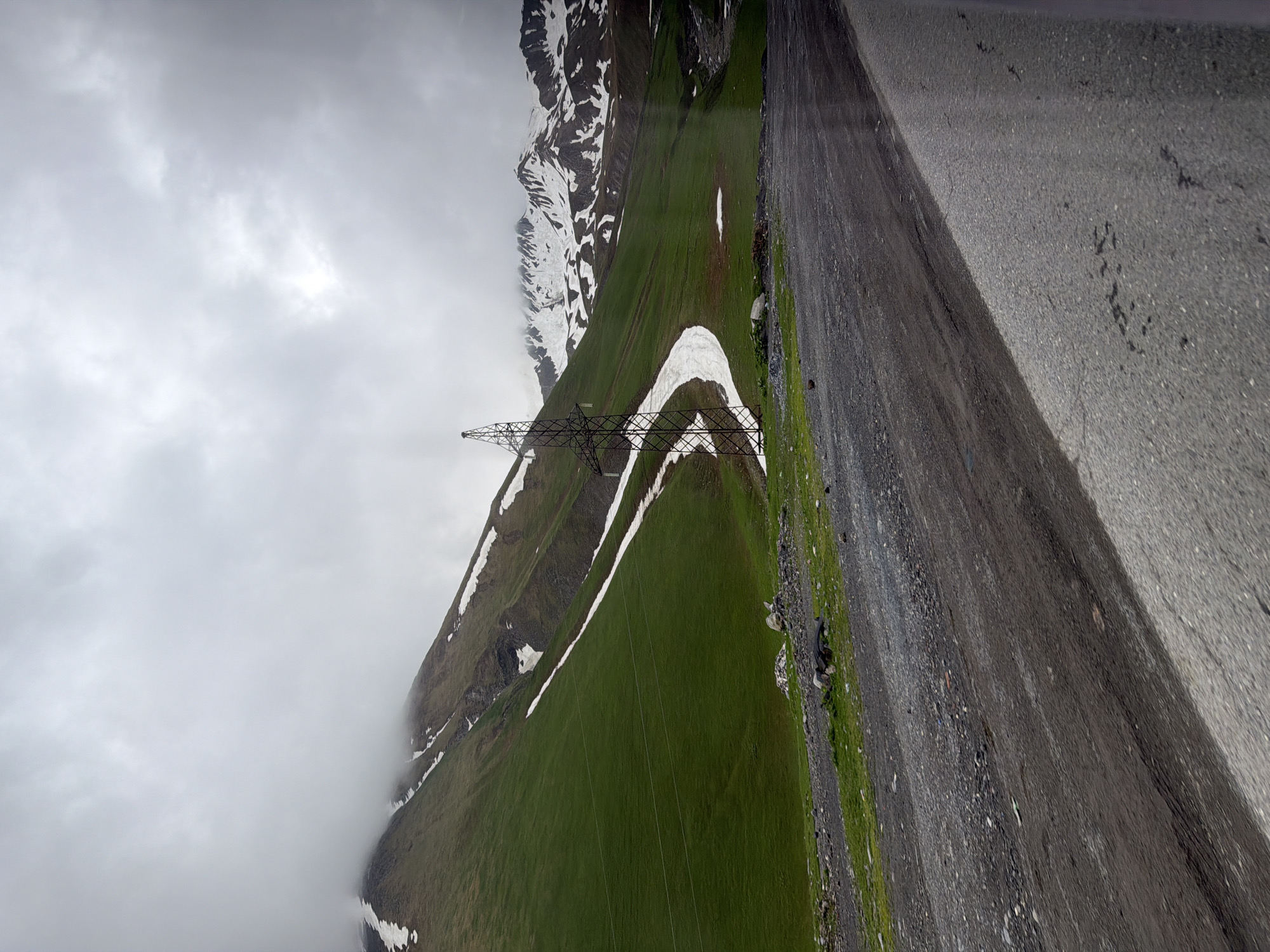 This is a landscape photograph taken from a tilted perspective, showing a mountainous region under a cloudy sky. The scene is dominated by a steep, grassy mountainside with patches of snow, particularly a distinctive white ribbon of snow winding down the green slope. In the foreground, a road made of gravel and asphalt runs diagonally across the frame, with a distinct shadow cast upon it. A tall, lattice-like electricity pylon stands prominently on the mountainside, connected by wires that extend out of frame. The sky is overcast with dramatic, white clouds, suggesting a cool or possibly damp day. The lighting is diffused, creating a soft contrast. The overall atmosphere is one of natural grandeur, with signs of human infrastructure in the form of the pylon and road. There is no visible text in the image.