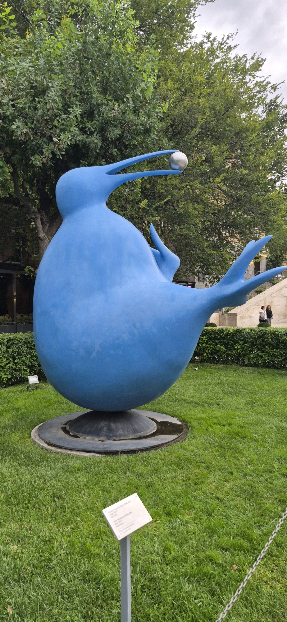 A large, bright blue abstract sculpture resembling a bird with a silver orb in its beak is prominently displayed on a patch of green grass in Yerevan, Armenia. The sculpture, situated on a circular dark base, features a plump body, a long, thin beak, and two wing-like appendages extending from its sides. Behind the sculpture, lush green trees form a dense backdrop, with a hint of a cloudy sky peeking through the foliage. In the middle ground, a neatly trimmed hedge separates the sculpture's area from a paved walkway and a stone staircase. Two individuals, a woman in a dark top and a lighter-colored bottom and another woman in a light-colored top and dark pants, are seen walking on the paved area near the staircase. A small white signpost with text in English is visible in the foreground on the grass, likely indicating information about the sculpture. The overall scene suggests an outdoor public art installation within a park or garden setting in Yerevan.