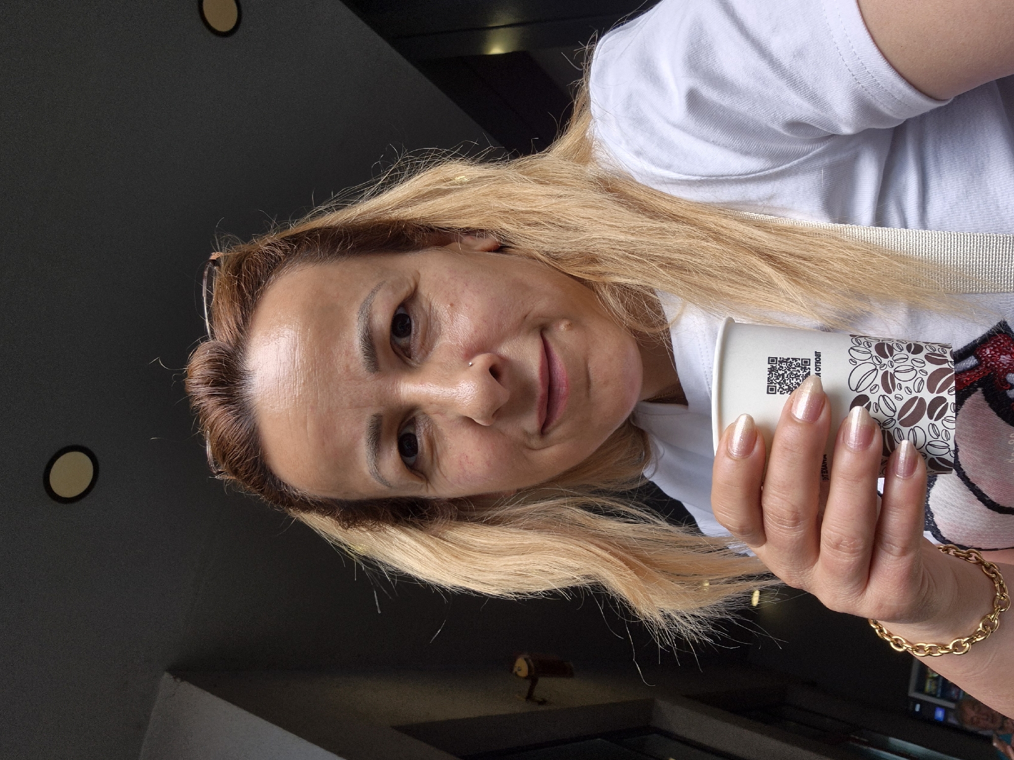A woman with blonde hair and a white t-shirt is holding a coffee cup in her left hand. She is looking at the camera and smiling. She is wearing a gold bracelet on her left wrist. The background is dark and appears to be an indoor or covered outdoor space with some lighting fixtures on the ceiling.
