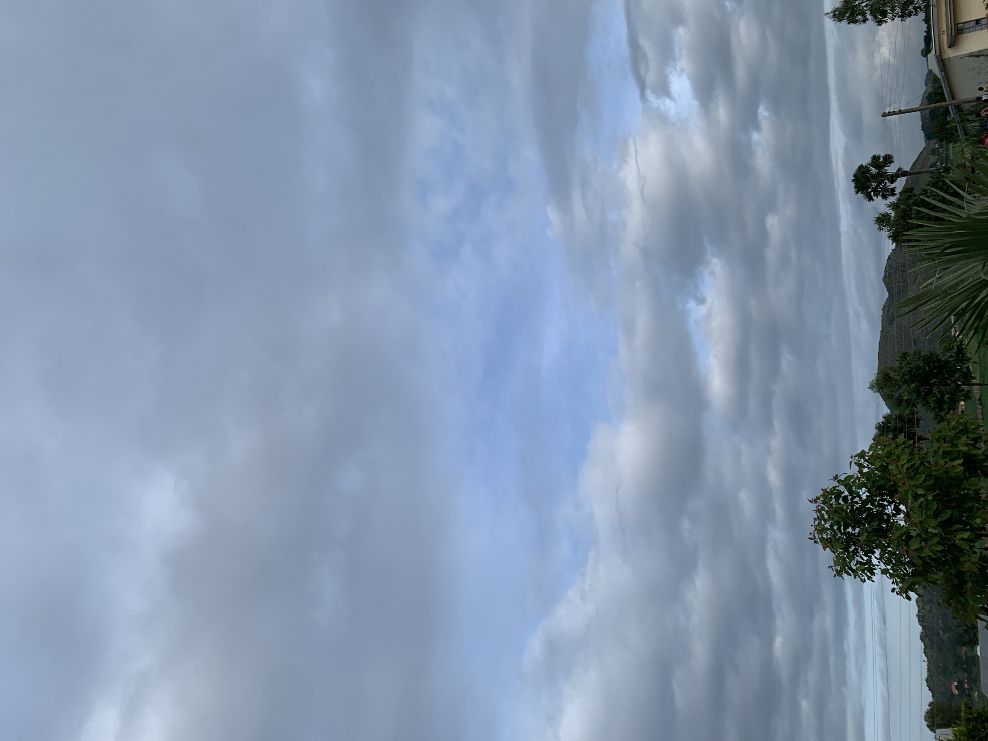 The image captures a cloudy sky over a landscape in Kuru Karama, Nigeria. The sky is filled with various shades of gray and blue clouds, with patches of brighter sky peeking through. In the foreground, several trees are visible, including a large leafy tree on the left and palm trees further back. The terrain appears hilly with some vegetation and scattered buildings in the distance. A few utility poles and lines are also present. The overall impression is of an overcast day in a rural or semi-rural setting.