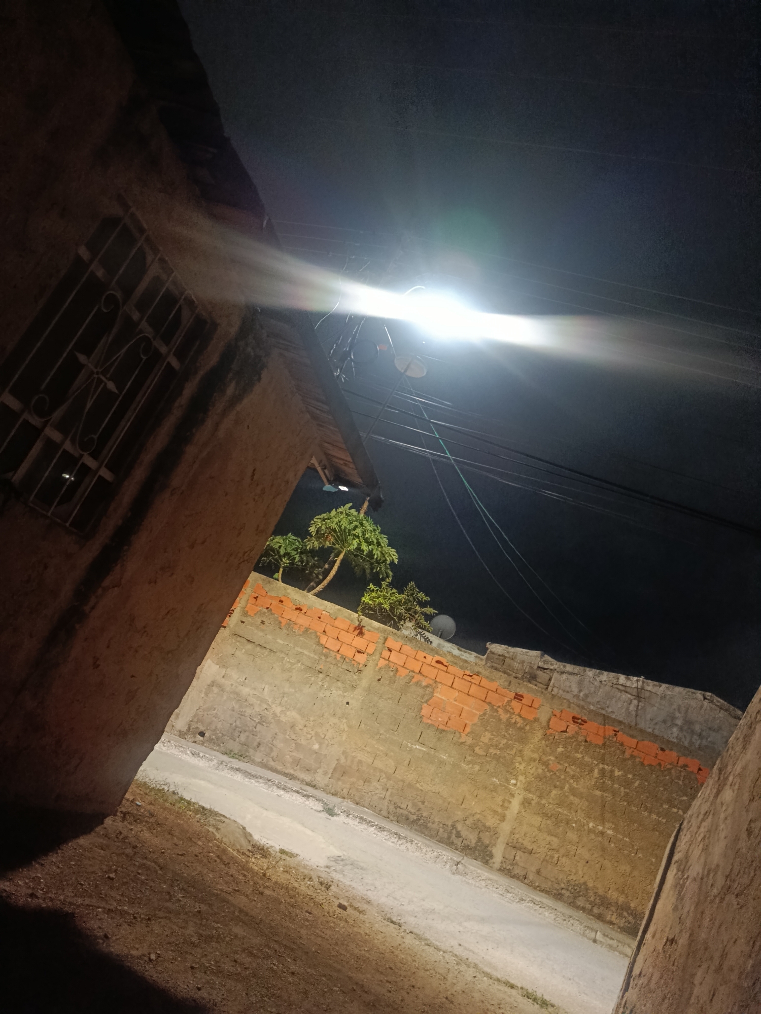 The image presents a night scene in a narrow alleyway, potentially located in the city of Juan Griego, Venezuela, though no immediate local cultural elements are visible. The alley is bounded by walls on either side. The wall on the right consists of irregular bricks, some of which appear to be painted or partially covered in red. The wall on the left is a tall, aged structure with a barred window visible near the top. The alley floor is a light-colored pavement or dirt path that leads into the distance. The overhead space is dominated by a bright street light, casting a strong glare and light beams in the darkness. Electrical wires are strung overhead. A tree is visible in the background. No people, interactions, or actions are explicitly evident in the image. The environment is primarily dark, punctuated by the artificial light source and the textures of the surrounding structures.
