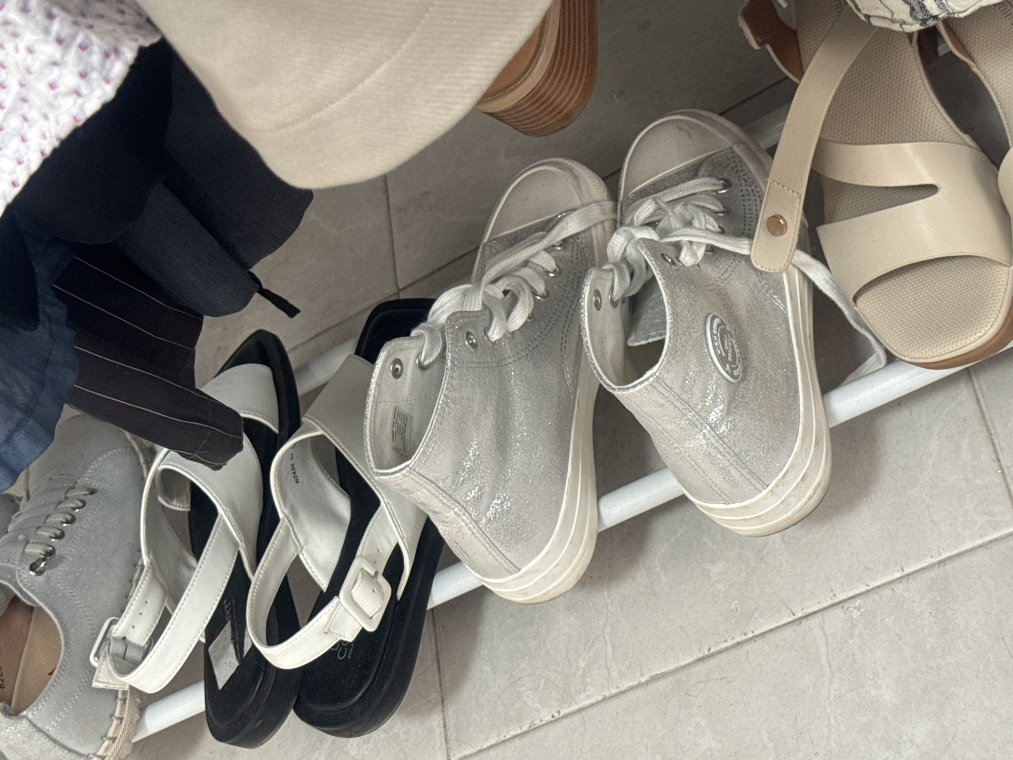 The image shows a variety of shoes on a white rack, likely in a closet. There are two pairs of silver high-top sneakers with white laces. To the left, there are two pairs of sandals: one pair with a white strap and a black sole, and another gray shoe with a tan sole. Some clothing items are visible hanging above the shoes, including a white and pink patterned garment and various shades of gray and black. The background consists of a tiled floor. The location is Ramat Gan, Israel, based on visual inspection.